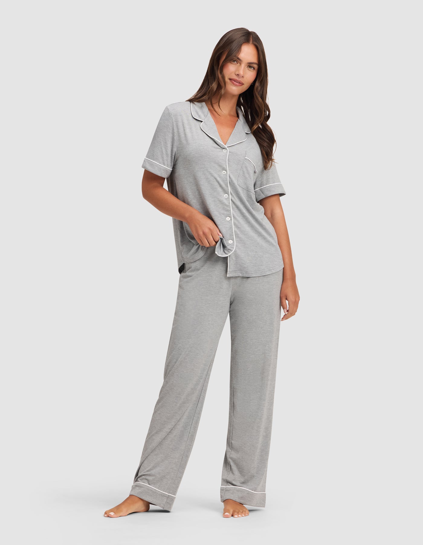 A woman stands barefoot in HIDE's Women's Bamboo Stretch-Knit Pajama Pant, paired with a matching light gray short-sleeve button-up top with white piping, posing relaxedly against a plain background.