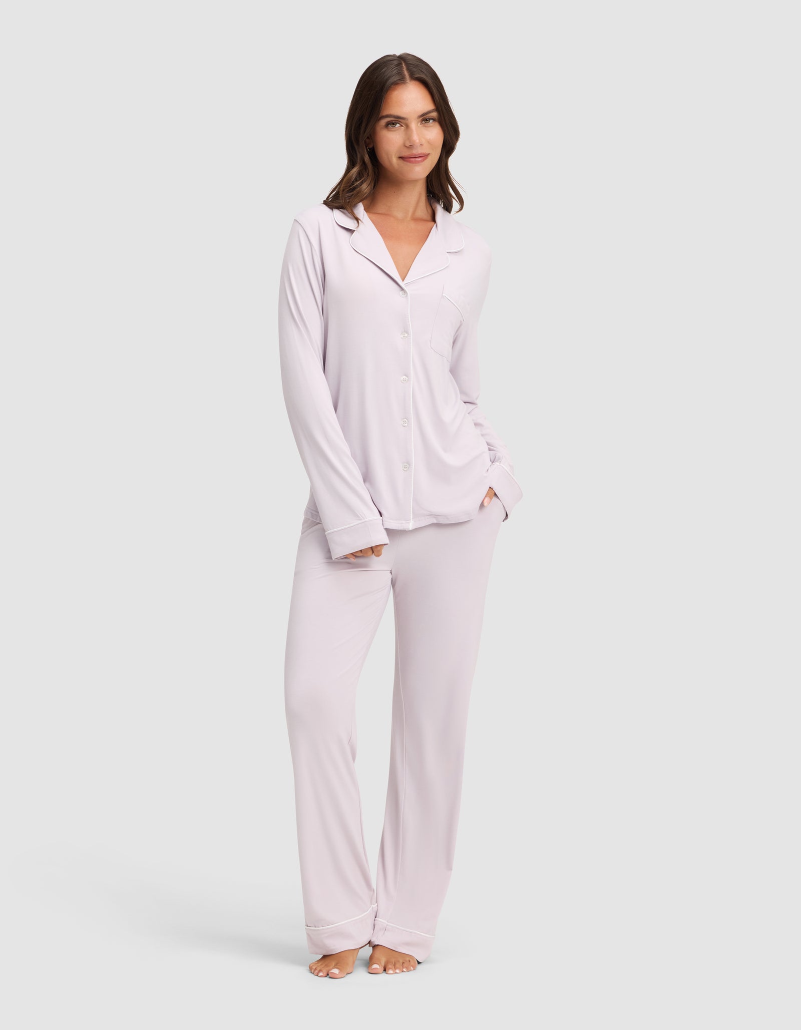 A woman wears HIDE Women's Bamboo Stretch-Knit Pajama Pants with a light-colored, long-sleeve button-up top. With one hand in her pocket, she smiles slightly against a plain light background.