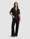 A woman stands barefoot against a light gray background, wearing Cozy Earth’s Women's Bamboo Stretch-Knit Short Sleeve & Pant Pajama Set in black with white piping. The set includes a short-sleeved button-up top and long pants. |Color:Black