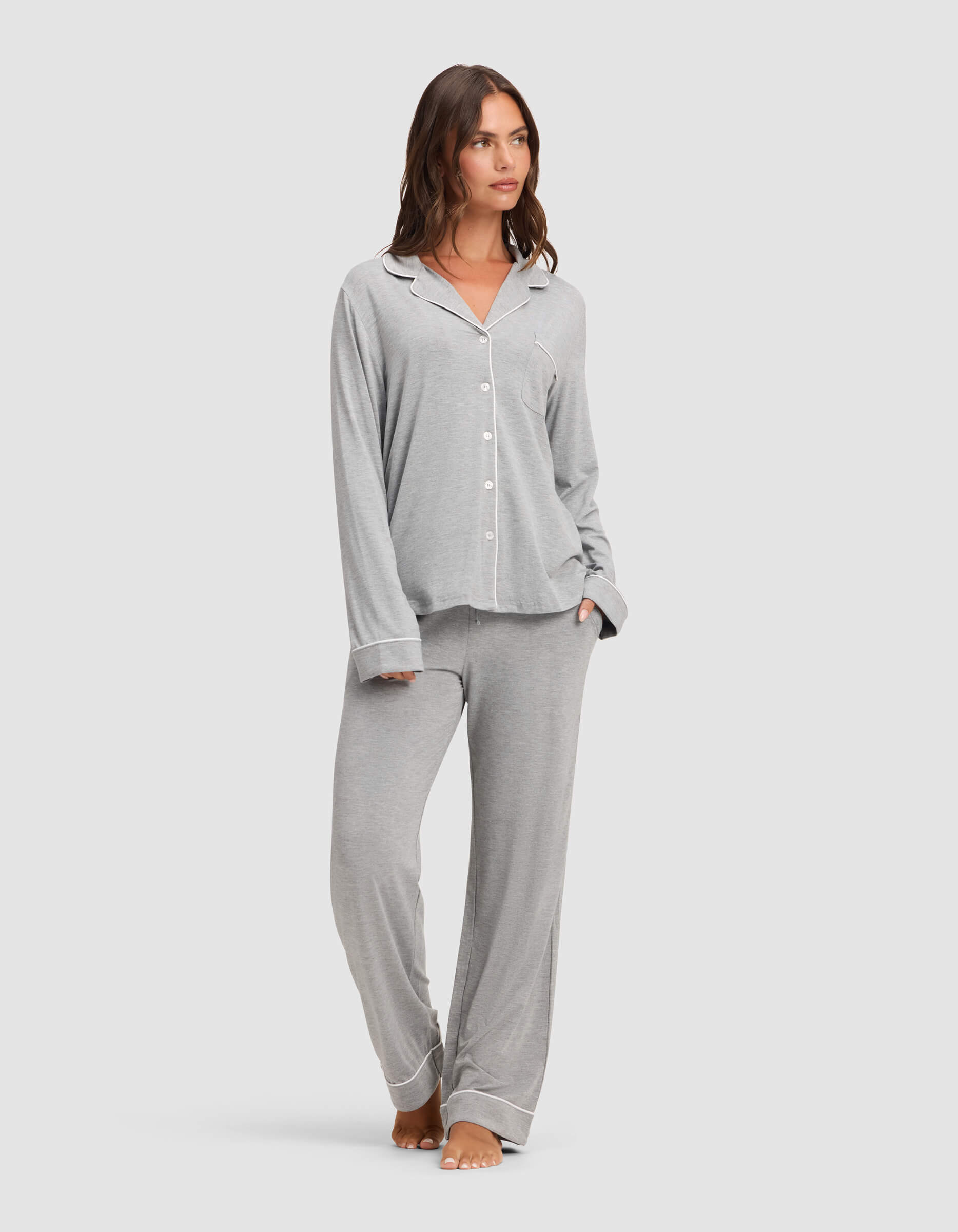 A woman stands barefoot against a plain light background, wearing the Cozy Earth Women's Bamboo Stretch-Knit Long Sleeve Pajama Set in light grey with white piping, featuring a button-up top and matching pants. |Color:Grey