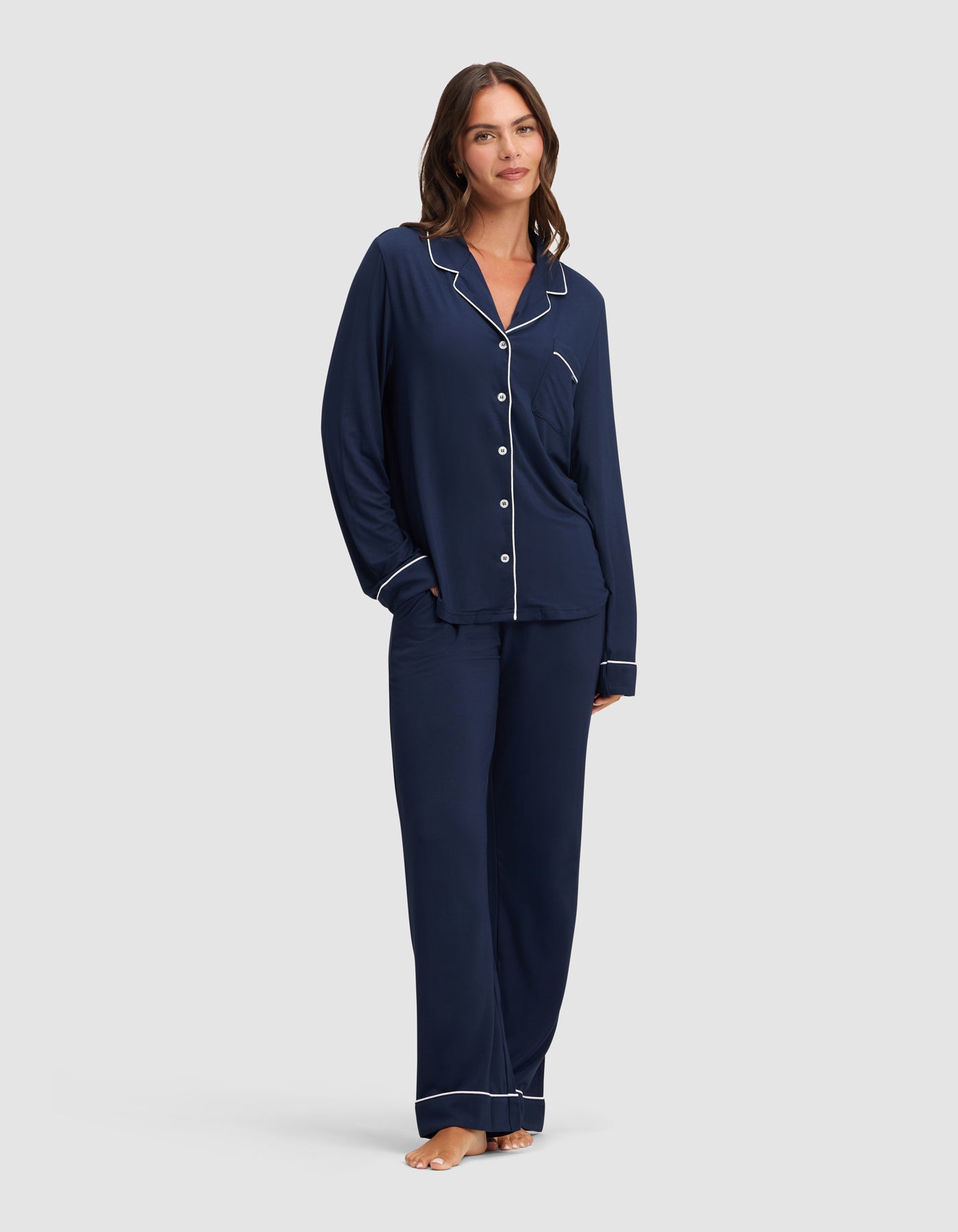 A woman poses barefoot against a plain light background, smiling in the Cozy Earth Women's Bamboo Stretch-Knit Long Sleeve Pajama Set—navy blue with white piping, featuring a button-up top and loose-fitting pants. |Color:Navy