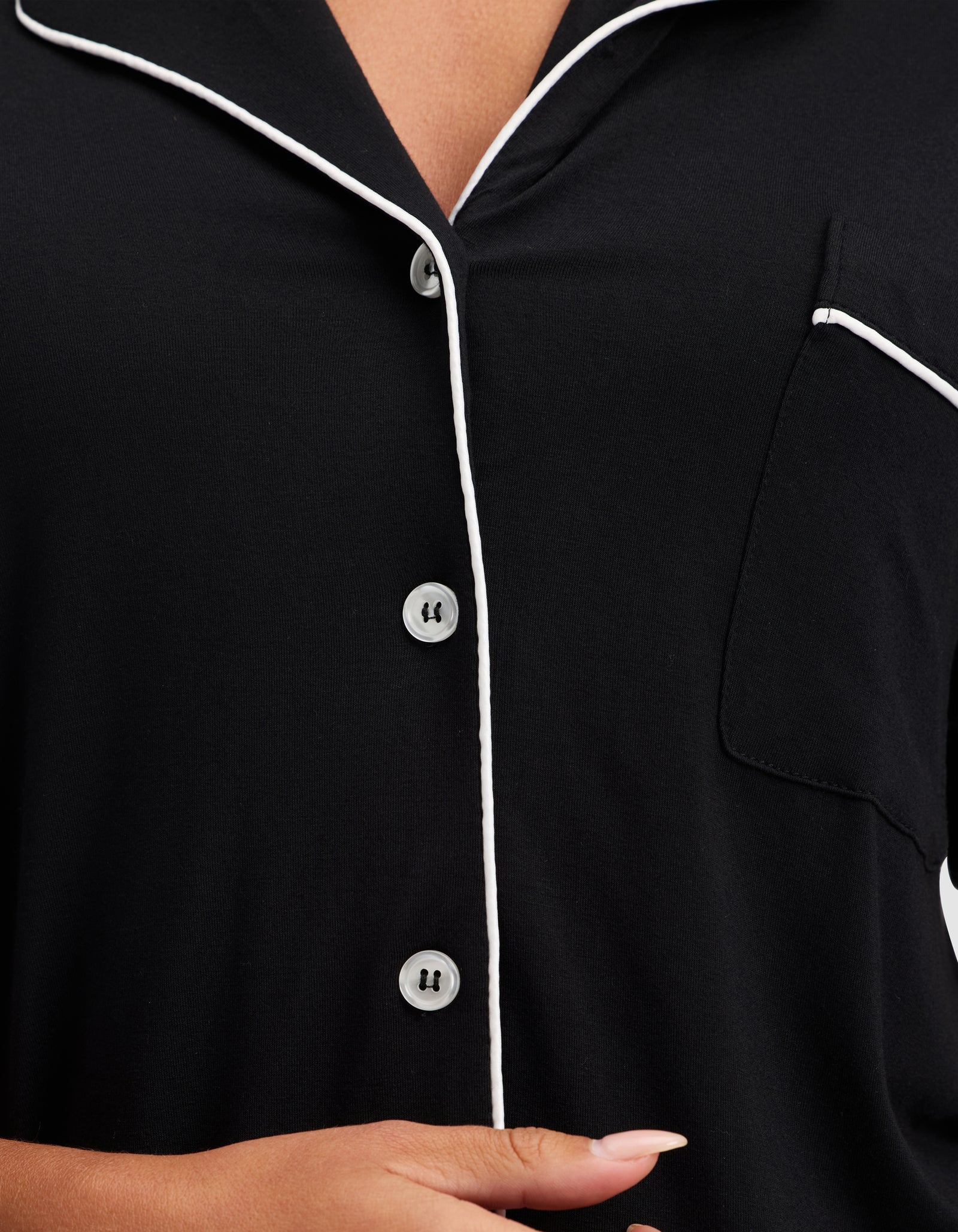 A close-up shows a person wearing the HIDE Women's Bamboo Stretch-Knit Short Sleeve Pajama Top in black, featuring white piping on the collar, button placket, and pocket edge, with their hand visible near the bottom of the frame.