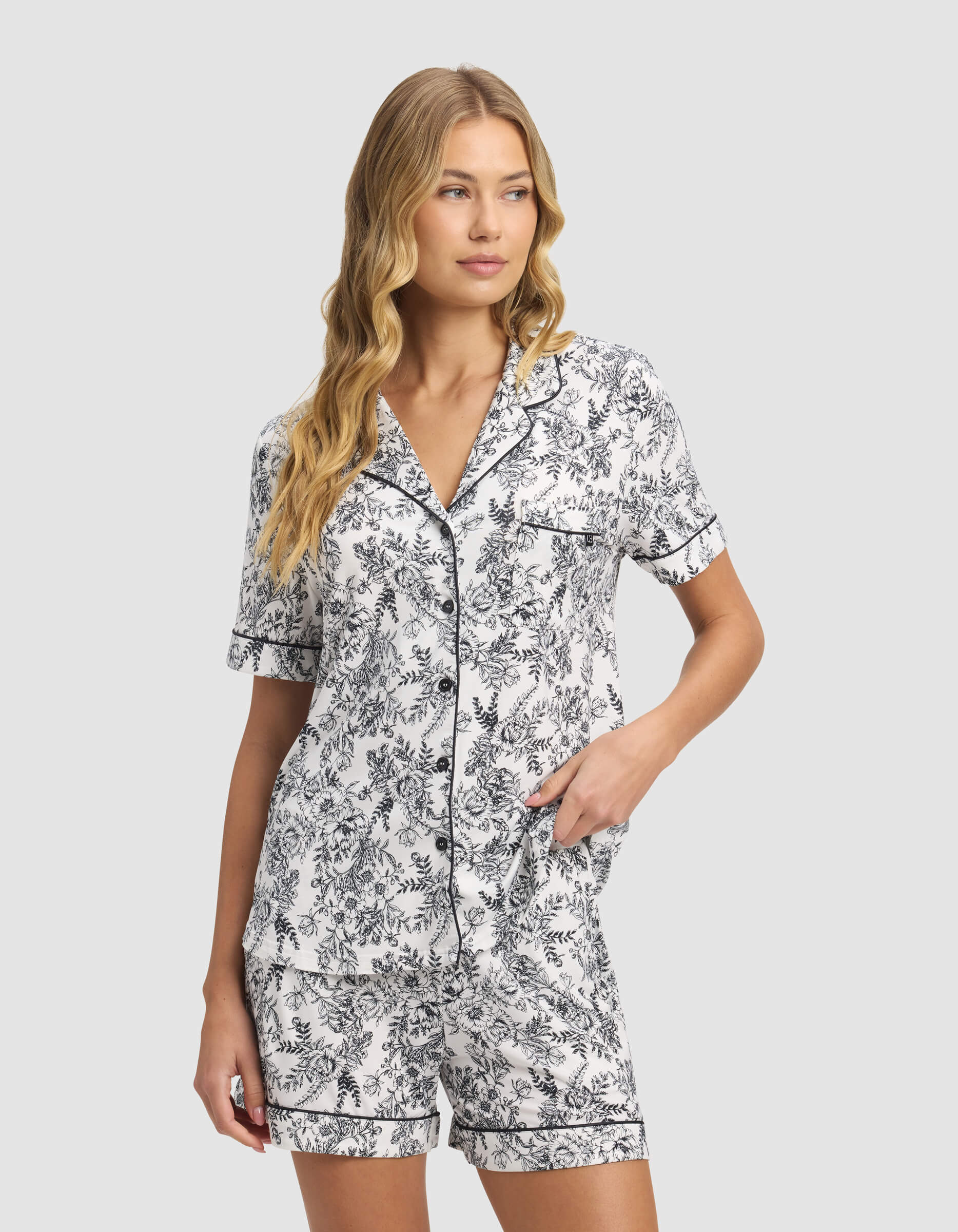A woman wears the Cozy Earth Women's Bamboo Stretch-Knit Short Sleeve Pajama Top, featuring a black and white floral pattern, standing with one hand in her pocket and looking to the side.|Color:Jet Black Toile