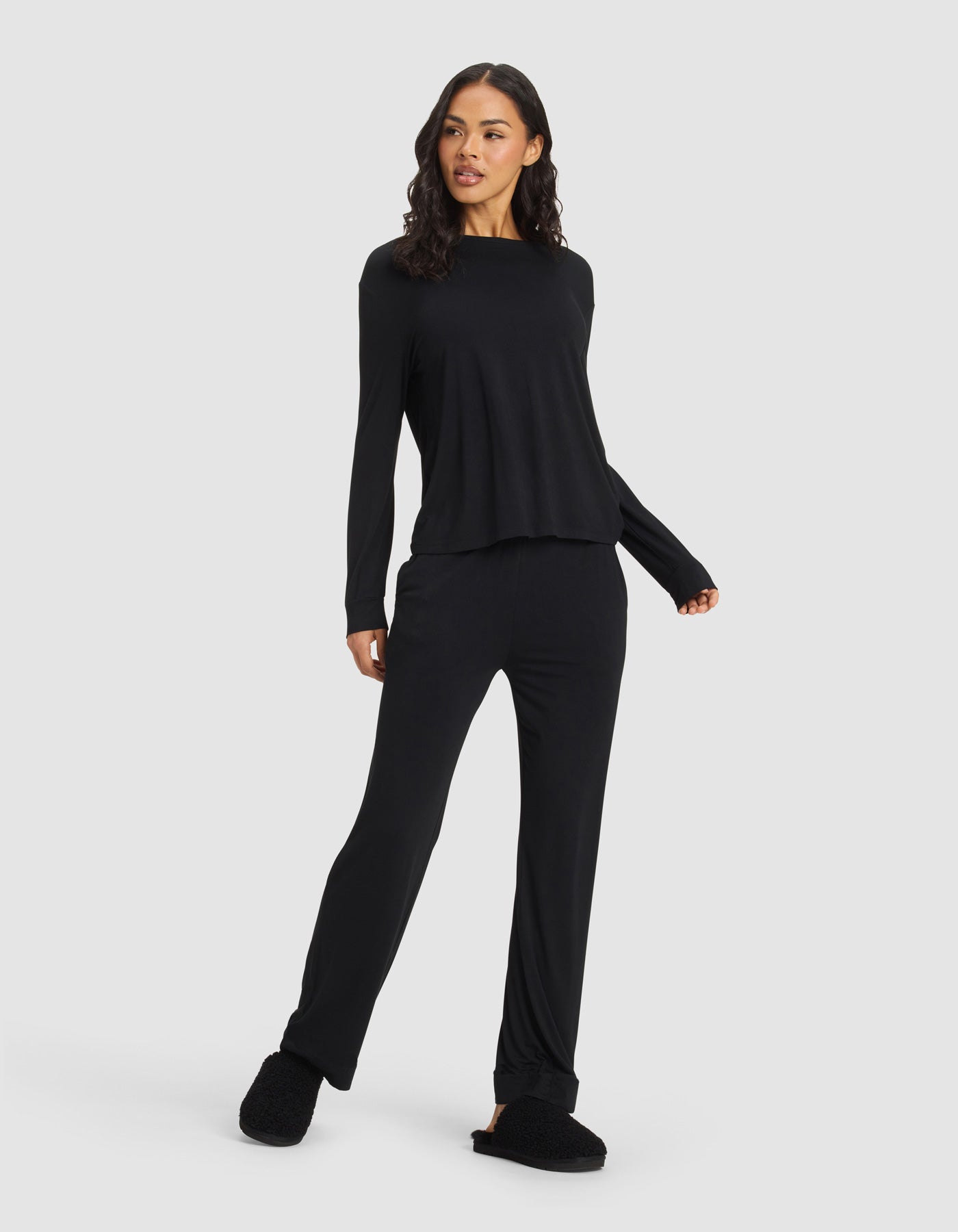 A woman wears the Cozy Earth Women's Bamboo Stretch-Knit Long Sleeve Lounge Tee & Pant Set in black, standing against a plain background with one hand on her hip and the other arm relaxed, looking to the side.|Color:Black