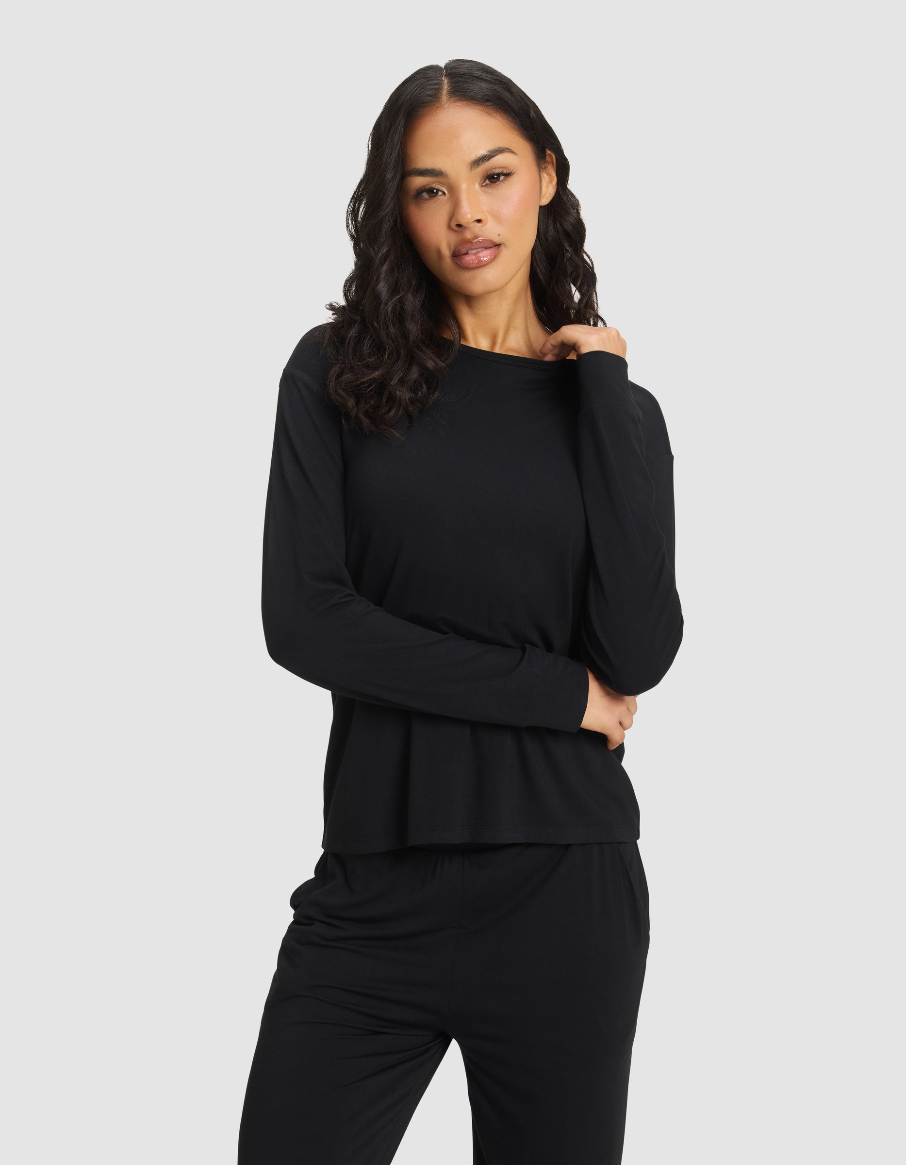 A woman with wavy dark hair models the Cozy Earth Women's Bamboo Stretch-Knit Long Sleeve Lounge Tee in black, standing against a plain light gray background with one hand on her shoulder and the other crossing her waist. |Color:Black