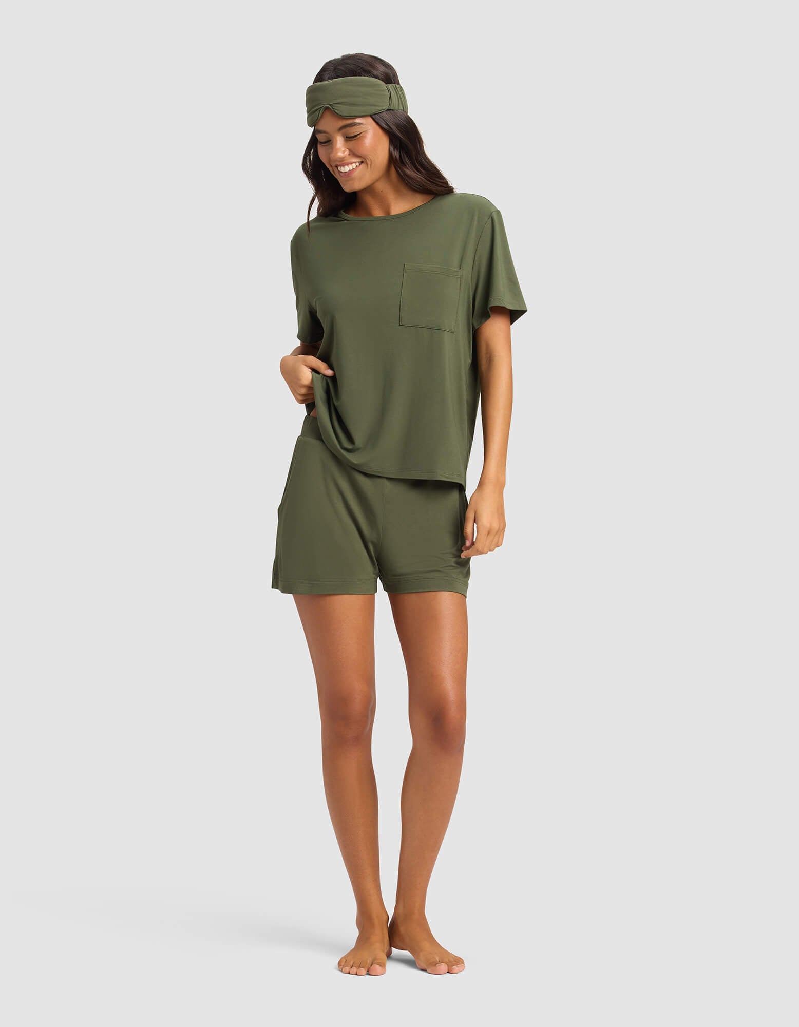 A woman stands barefoot and smiling, wearing Cozy Earth's Women's Bamboo Stretch-Knit Lounge Tee & Short Set in olive green with a matching headband, against a plain light background.|Color:Olive
