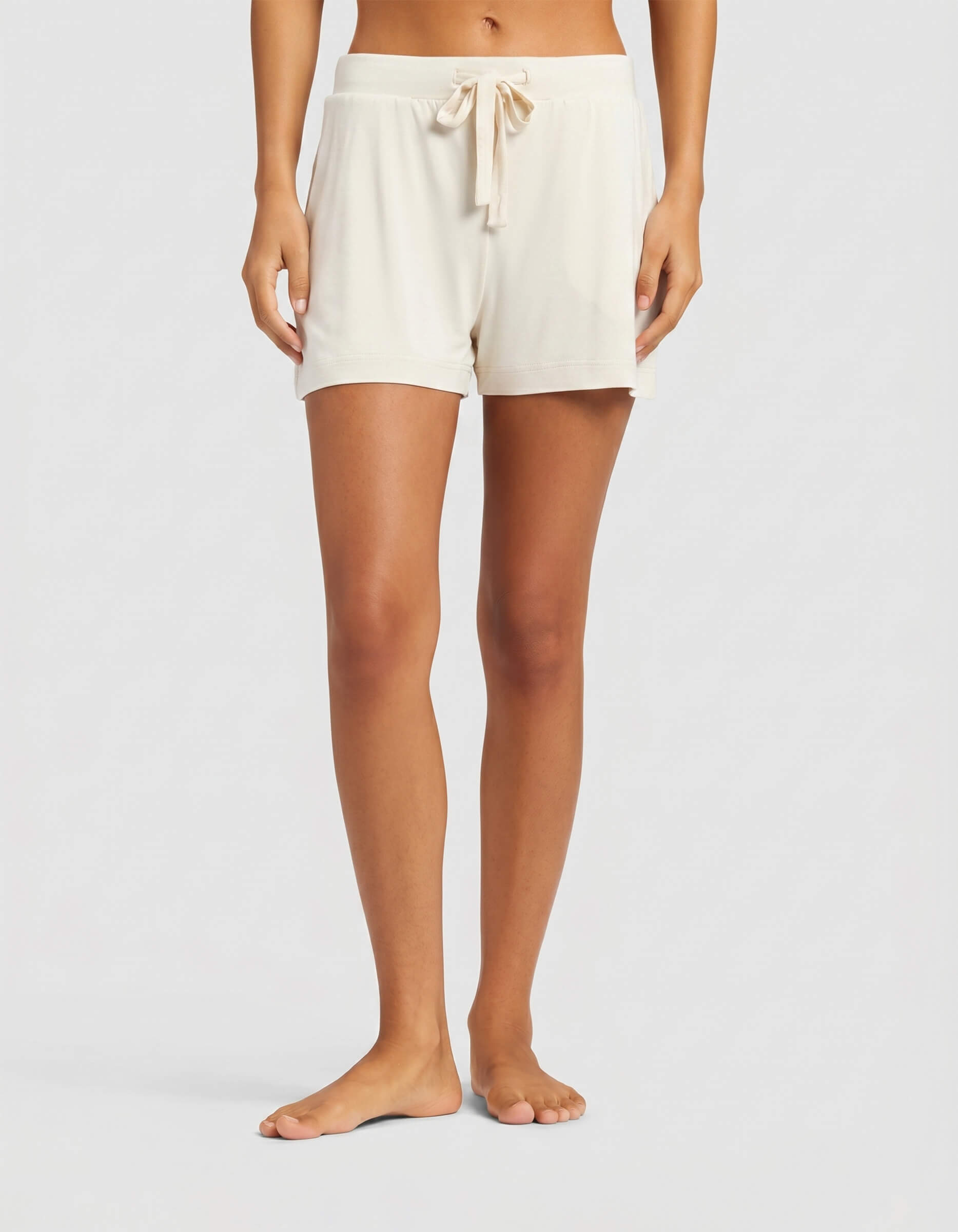 A woman stands barefoot against a plain light background, wearing Cozy Earth's Women's Bamboo Stretch-Knit Shorts in creme. The shorts feature a drawstring waist, relaxed fit, and side pockets.|Color:Ivory
