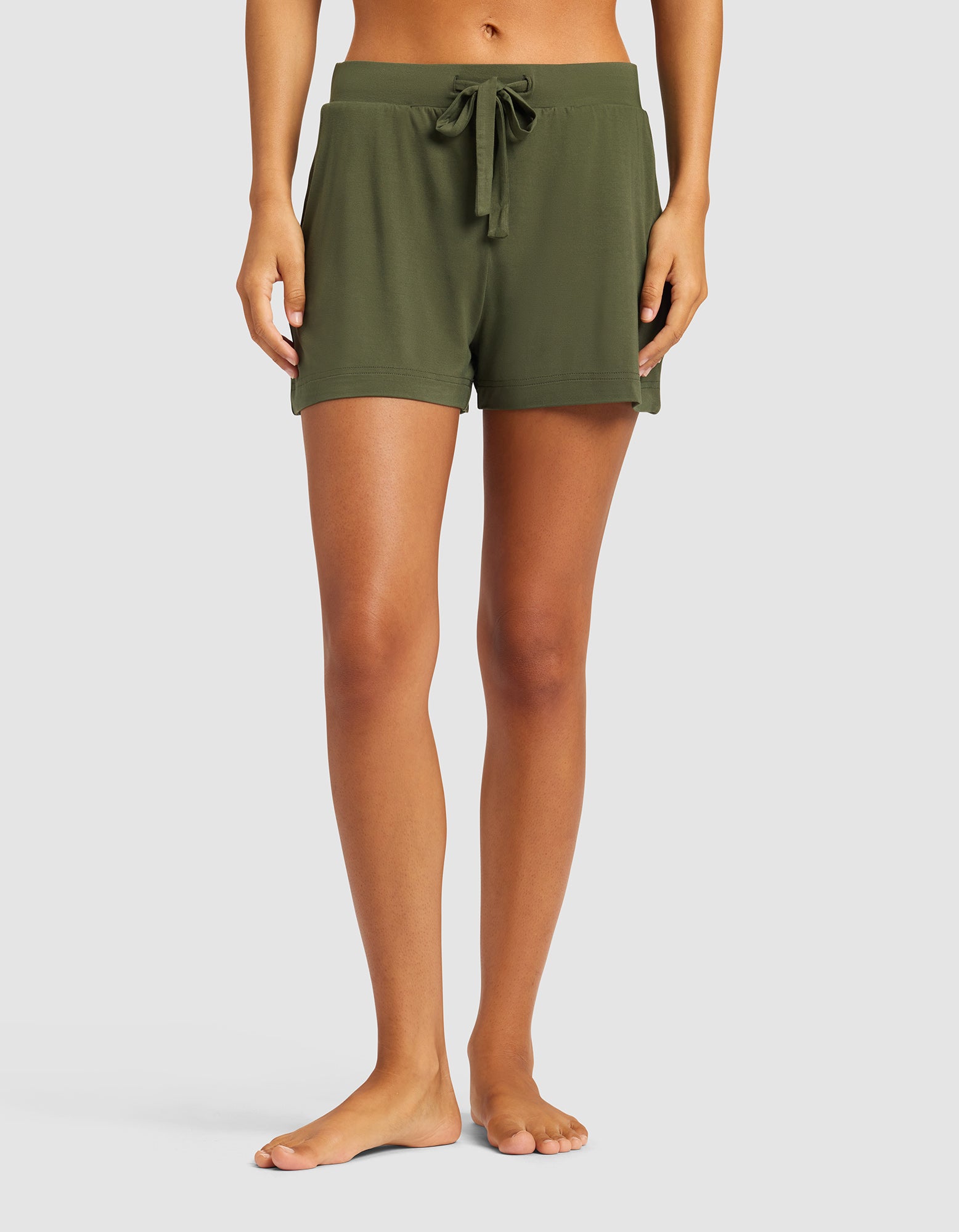 A woman stands barefoot against a plain light background, wearing Cozy Earth's Women's Bamboo Stretch-Knit Shorts in olive green. The shorts feature a drawstring waist, relaxed fit, and side pockets.|Color:Olive