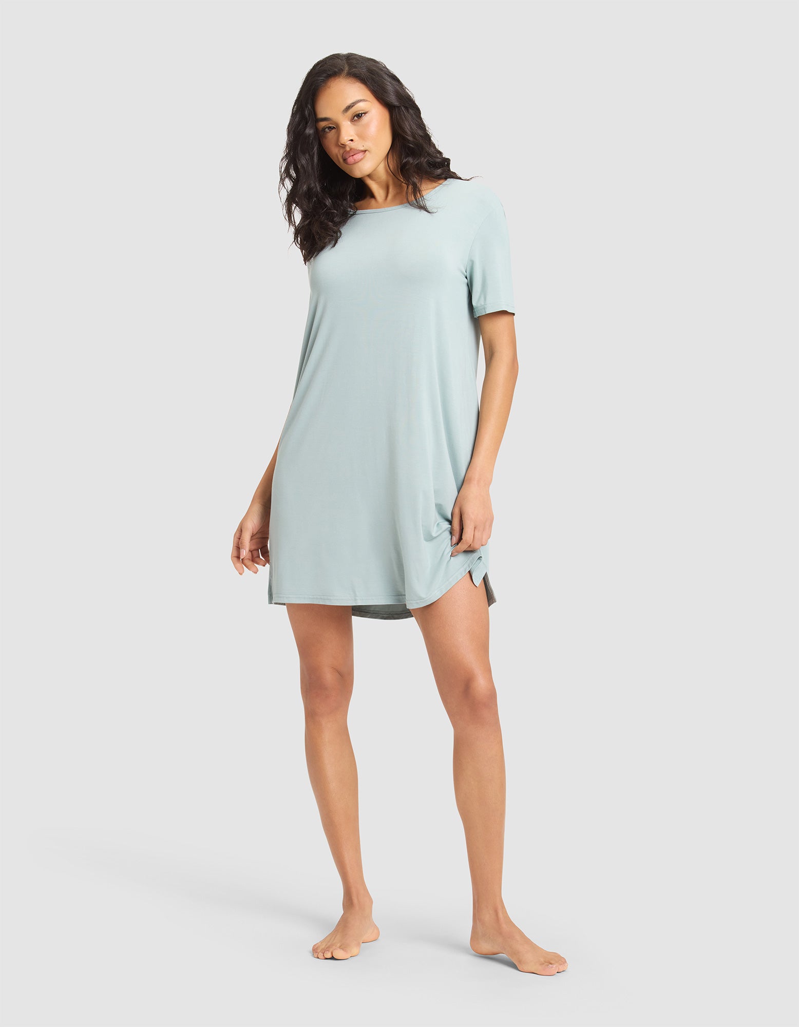 A woman stands barefoot wearing Cozy Earth's Women's Bamboo Stretch-Knit Sleep Dress, a relaxed-fit, light blue short-sleeve dress with a slight side slit, against a plain light gray background. |Color:Brook