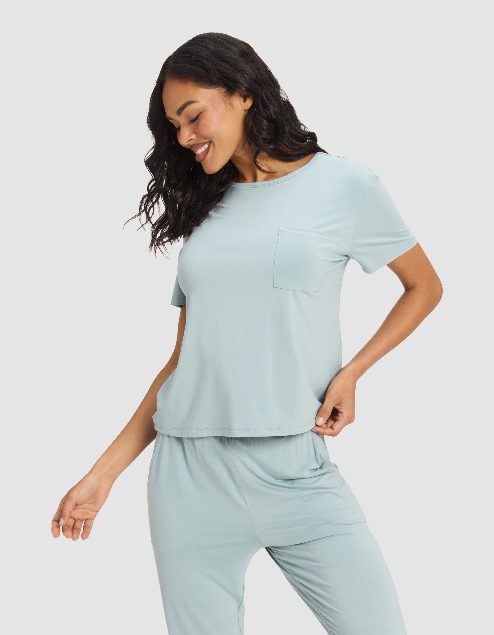 A woman smiles and looks down, wearing the Cozy Earth Women's Bamboo Stretch-Knit Lounge Tee in light blue, featuring short sleeves and a chest pocket, with one hand on her hip against a plain light background.|Color:Brook