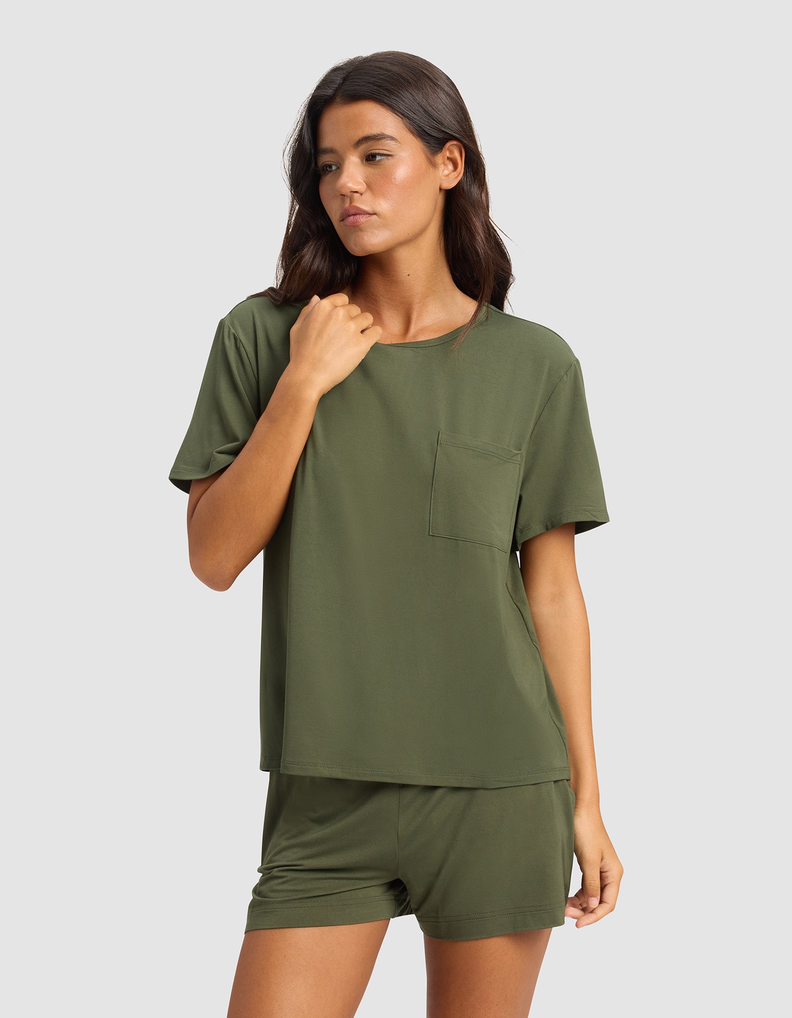 A woman with long brown hair wears the Cozy Earth Women's Bamboo Stretch-Knit Lounge Tee in olive green, paired with matching shorts, posing against a plain light gray background.|Color:Olive