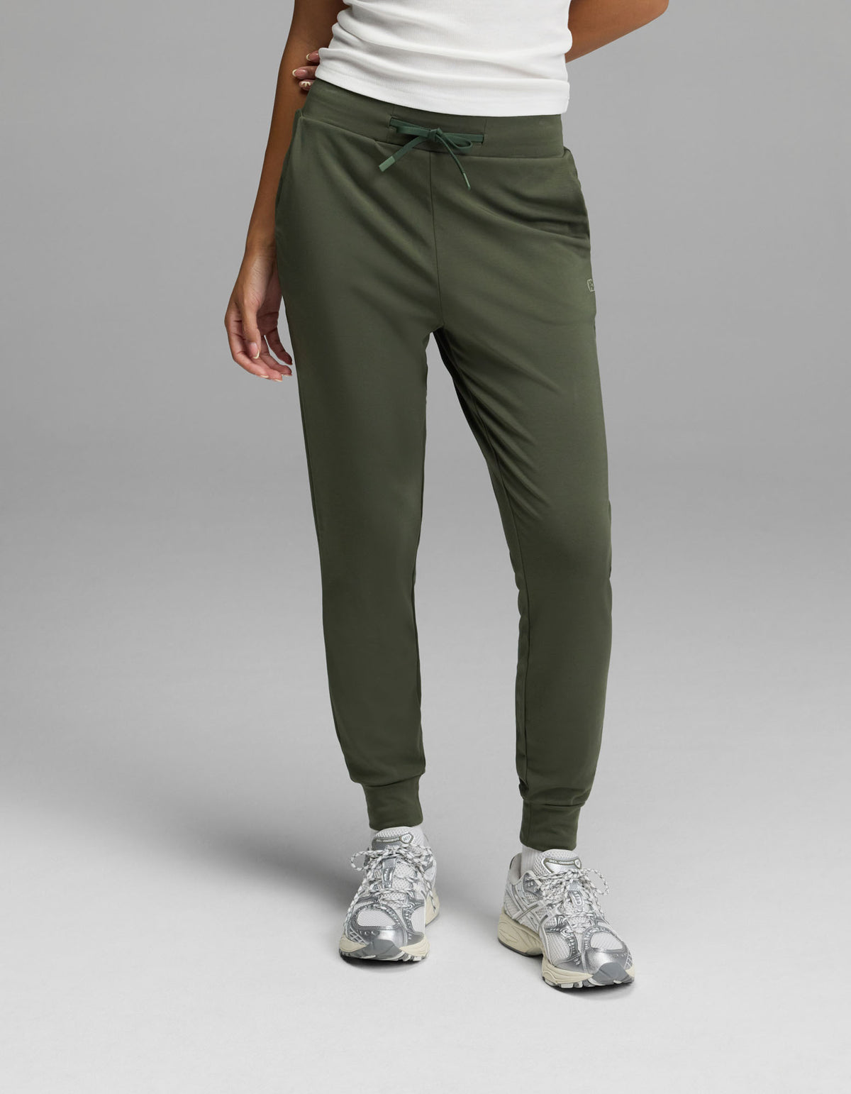 A person is shown from the waist down, wearing Cozy Earth’s Women’s Brushed Bamboo Jogger in olive green with a white top and silver sneakers, standing against a plain gray background. Only the lower body and part of one arm are visible.|Color:Olive