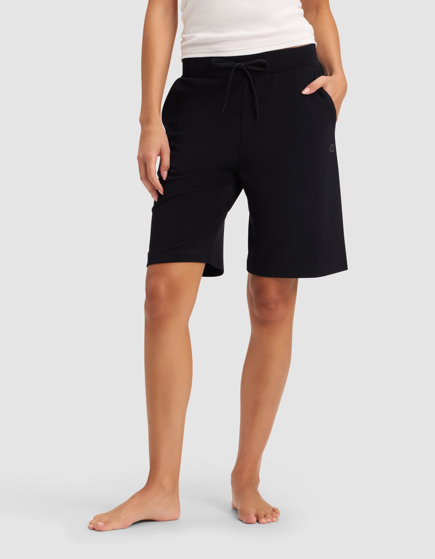 A woman models Cozy Earth’s Women’s Brushed Bamboo Bermuda Shorts, featuring a drawstring waistband in black, paired with a white sleeveless top, barefoot against a plain light gray background.|Color:Black