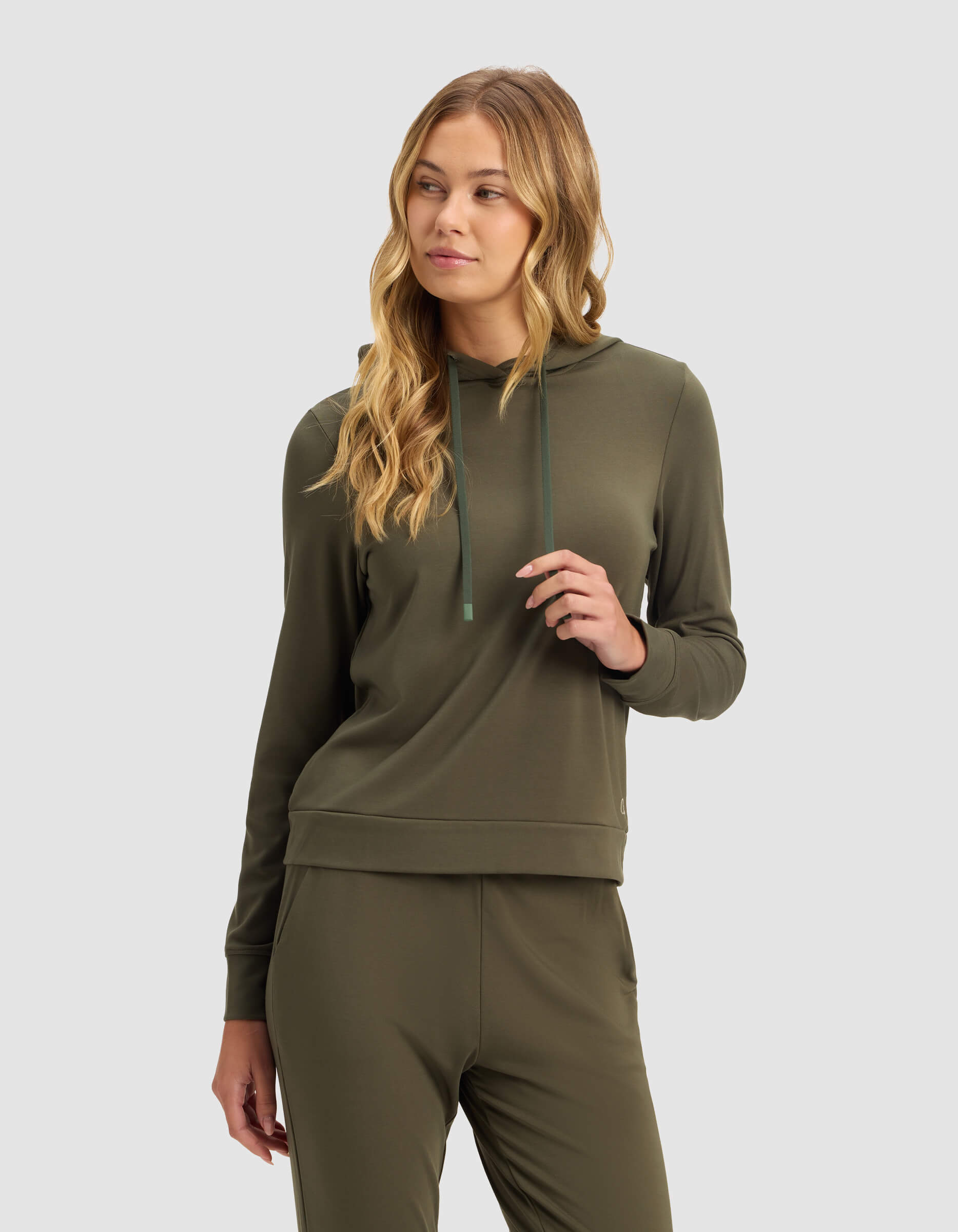 A woman with long blonde hair models the Cozy Earth Women's Brushed Bamboo Hoodie in Olive, standing against a plain light gray background and looking slightly left with a relaxed expression.|Color:Olive