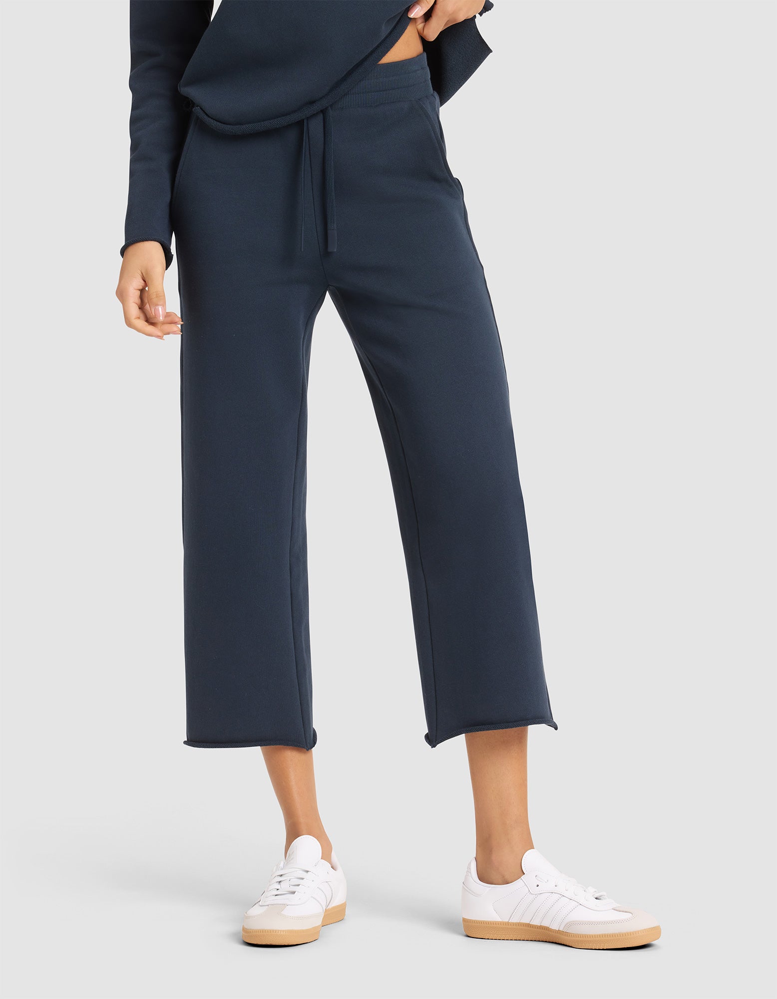 A woman stands against a plain light gray background wearing Cozy Earth's Women's Chelsea Cropped Pant in navy blue with a matching top and white sneakers. |Color:Eclipse