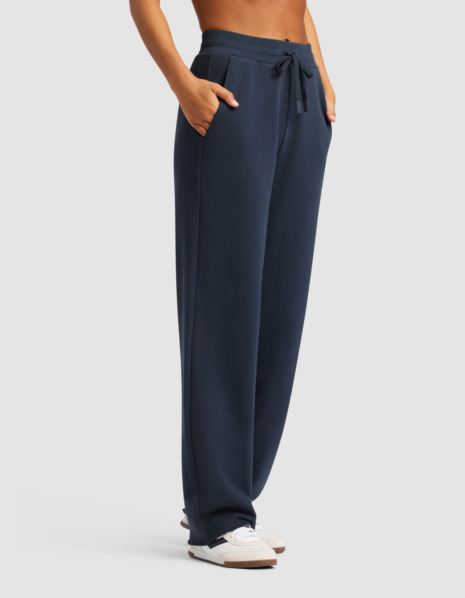 Someone wearing Cozy Earth's Chelsea Pant, featuring a loose fit and drawstring waist in navy blue, with hands in pockets and white sneakers, stands against a plain light background.