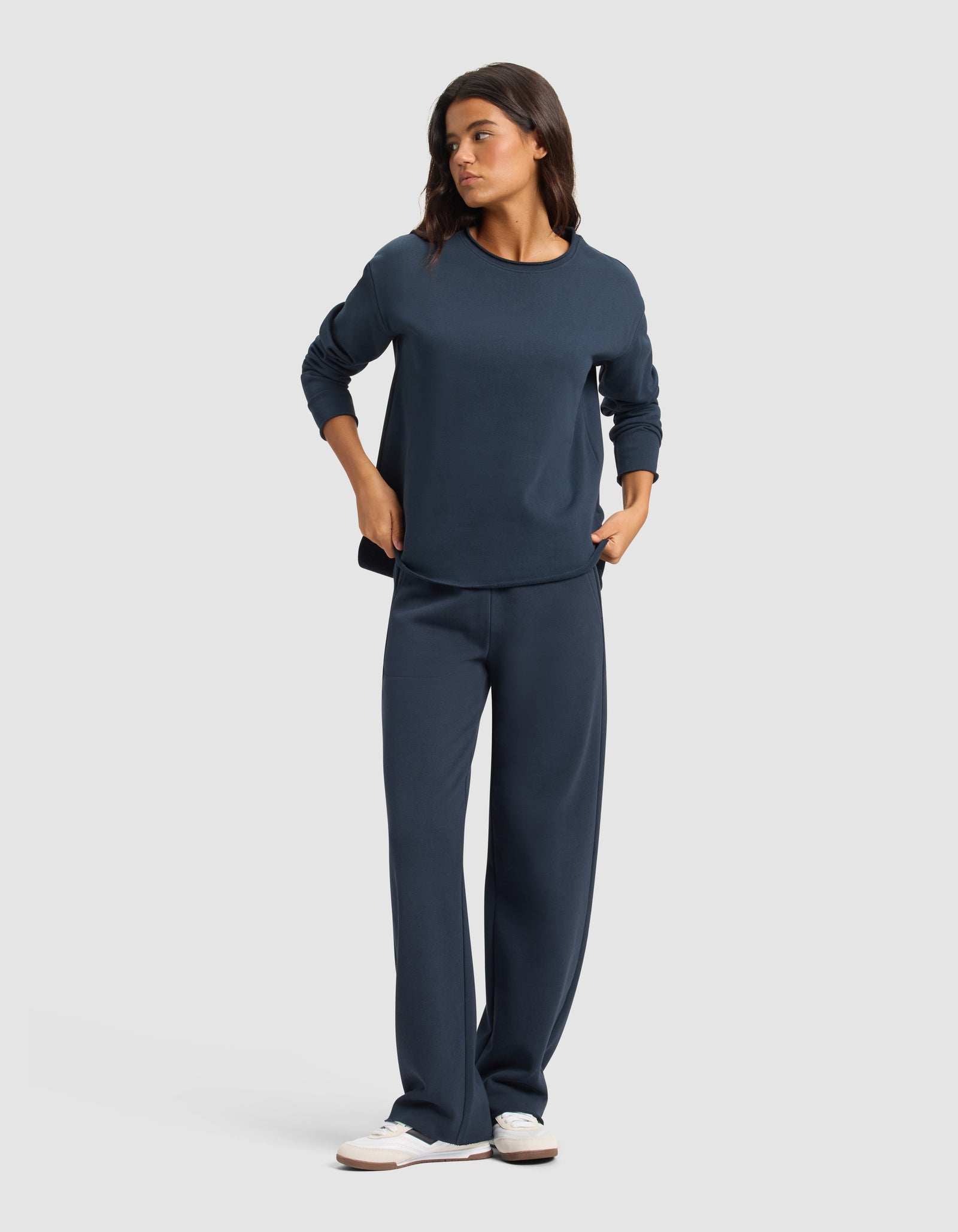 A woman stands against a plain background wearing Cozy Earth's Chelsea Pant in navy blue with a matching long-sleeve top and white sneakers. She has her hands in her pockets and looks to the side.