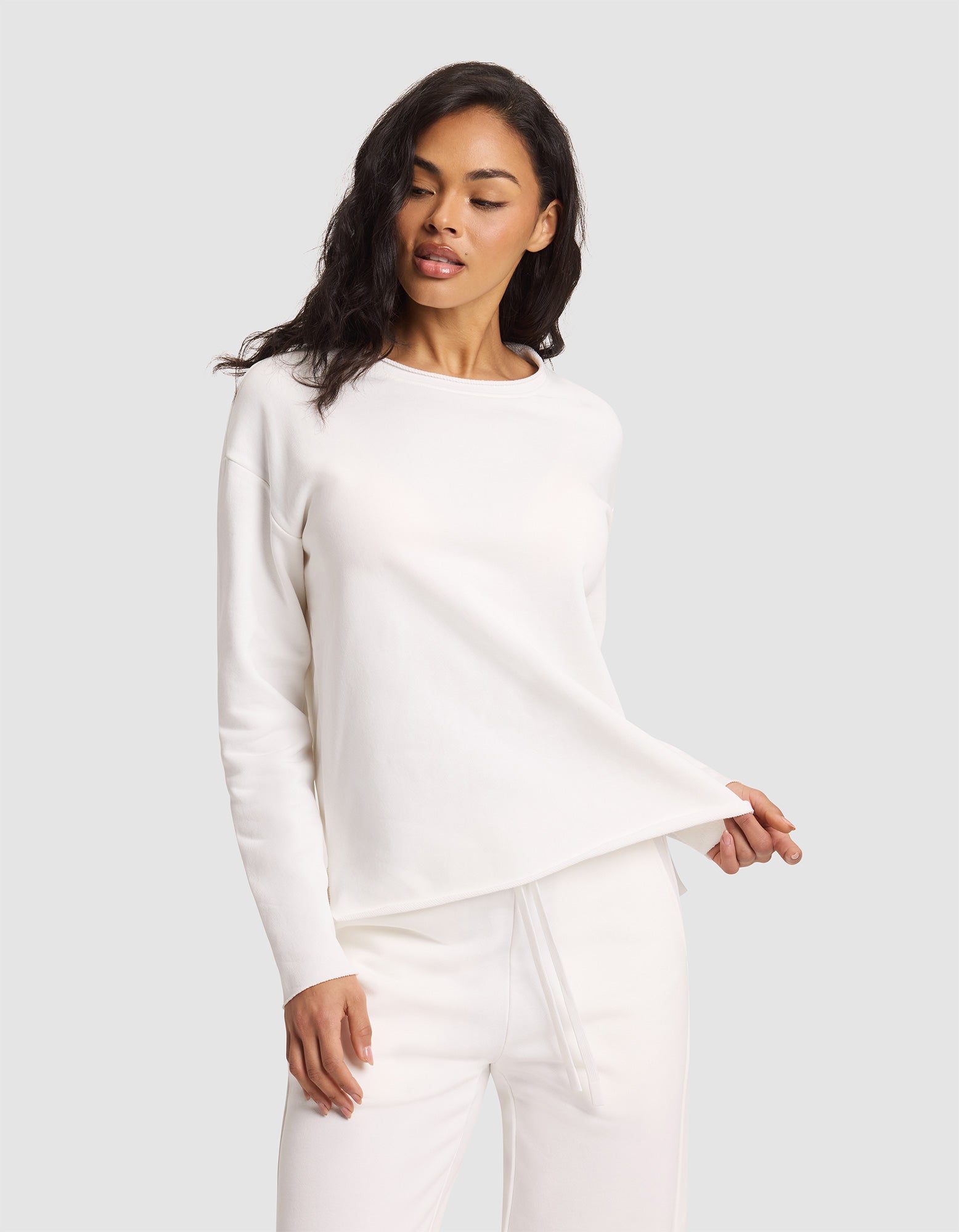 A woman poses against a light gray background wearing the Cozy Earth Women's Chelsea Pullover and matching pants in all white, holding the hem of the top with one hand while looking slightly downward. |Color:Bone