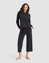 A woman with wavy dark hair models the Cozy Earth Women's Chelsea Set—matching black long-sleeve top and wide-leg cropped pants—standing barefoot with hands in her pockets against a plain light gray background. |Color:Jet Black