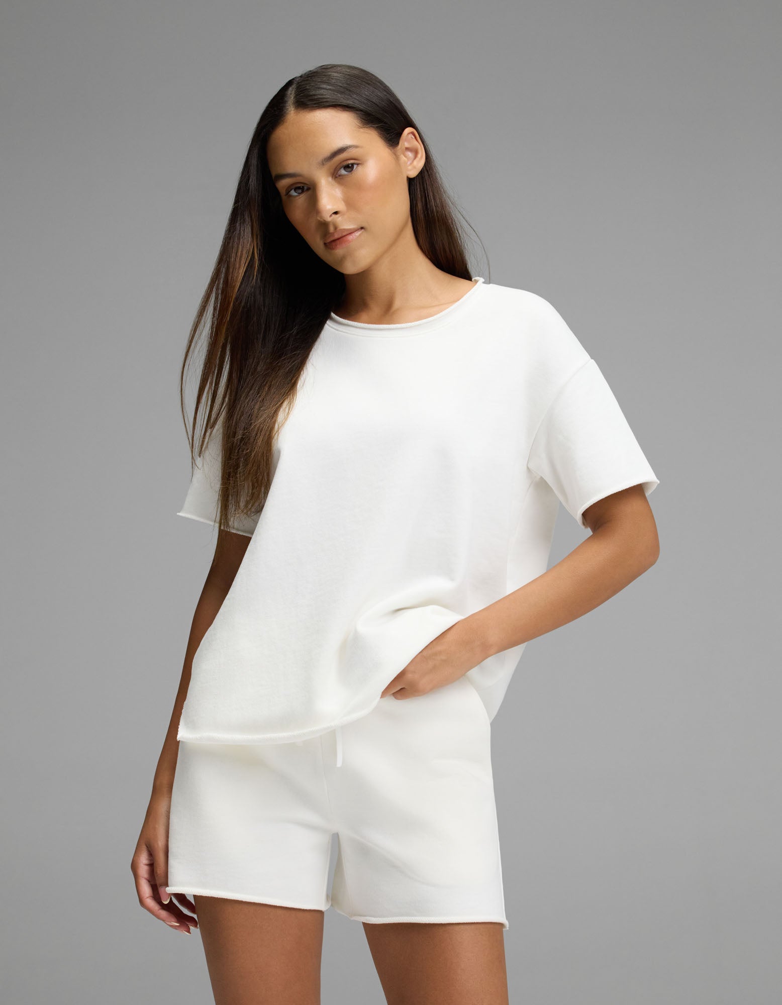 A woman with long, straight hair wears the women’s Chelsea Short Sleeve Set in Bone by Cozy Earth, standing against a plain gray background with one hand in her pocket.|Color:Bone