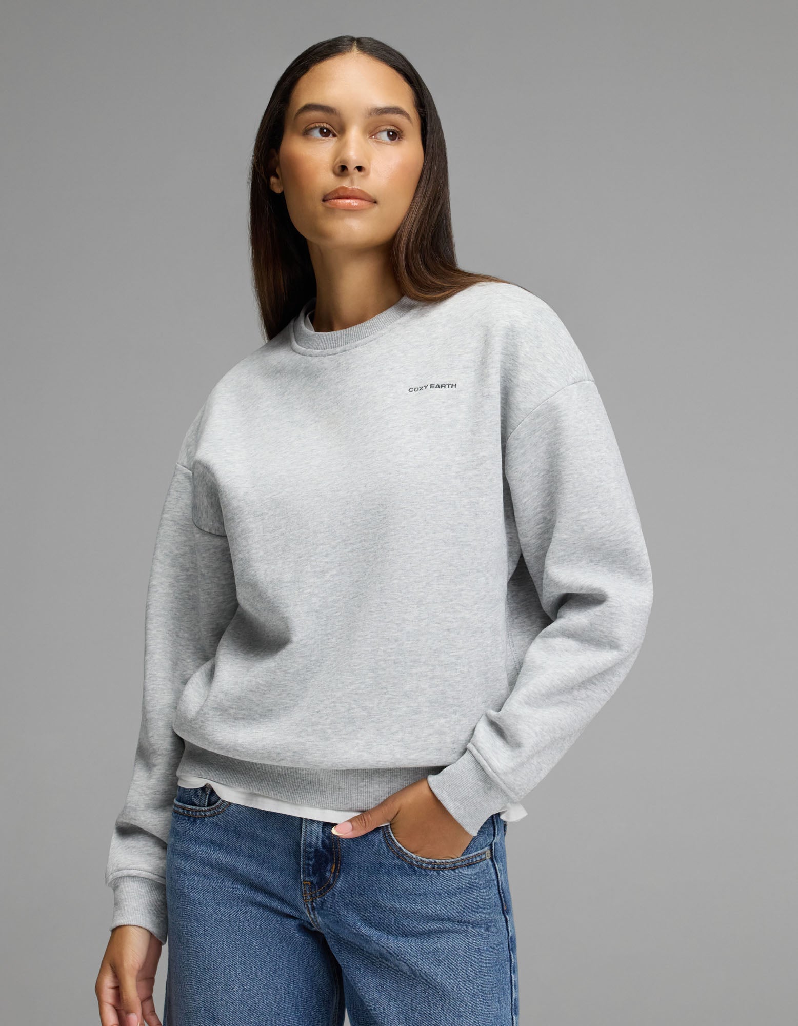 A person models the Cozy Earth Women’s Cityscape 2.0 Crewneck in Heather Grey, paired with blue jeans, standing against a plain gray background and glancing to the side with one hand in their pocket.|Color:Heather Grey