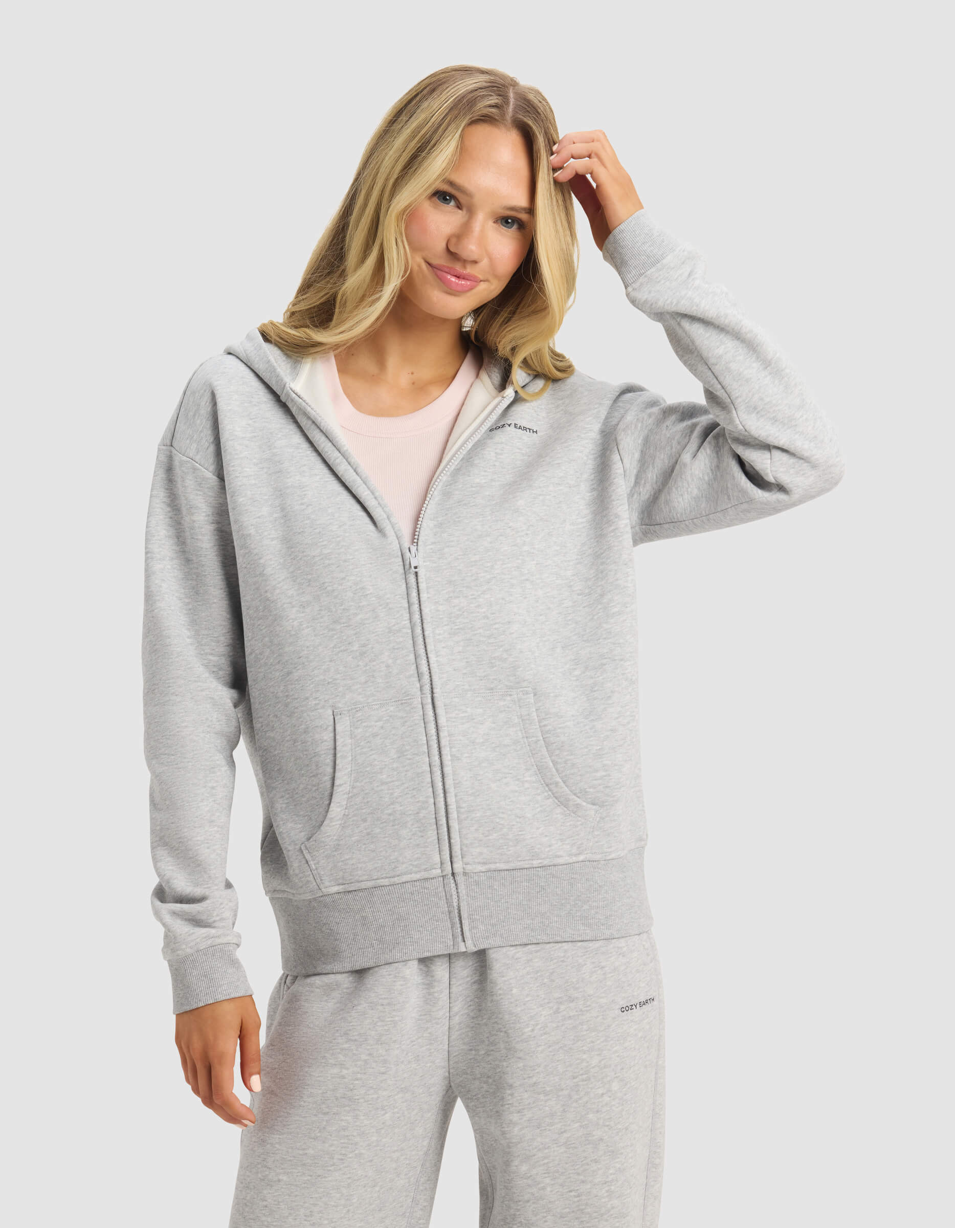 A woman with long blonde hair smiles and touches her hair while wearing Cozy Earth's Women's CityScape 2.0 Full Zip hoodie and matching sweatpants, standing against a plain light background.|Color:Heather Grey