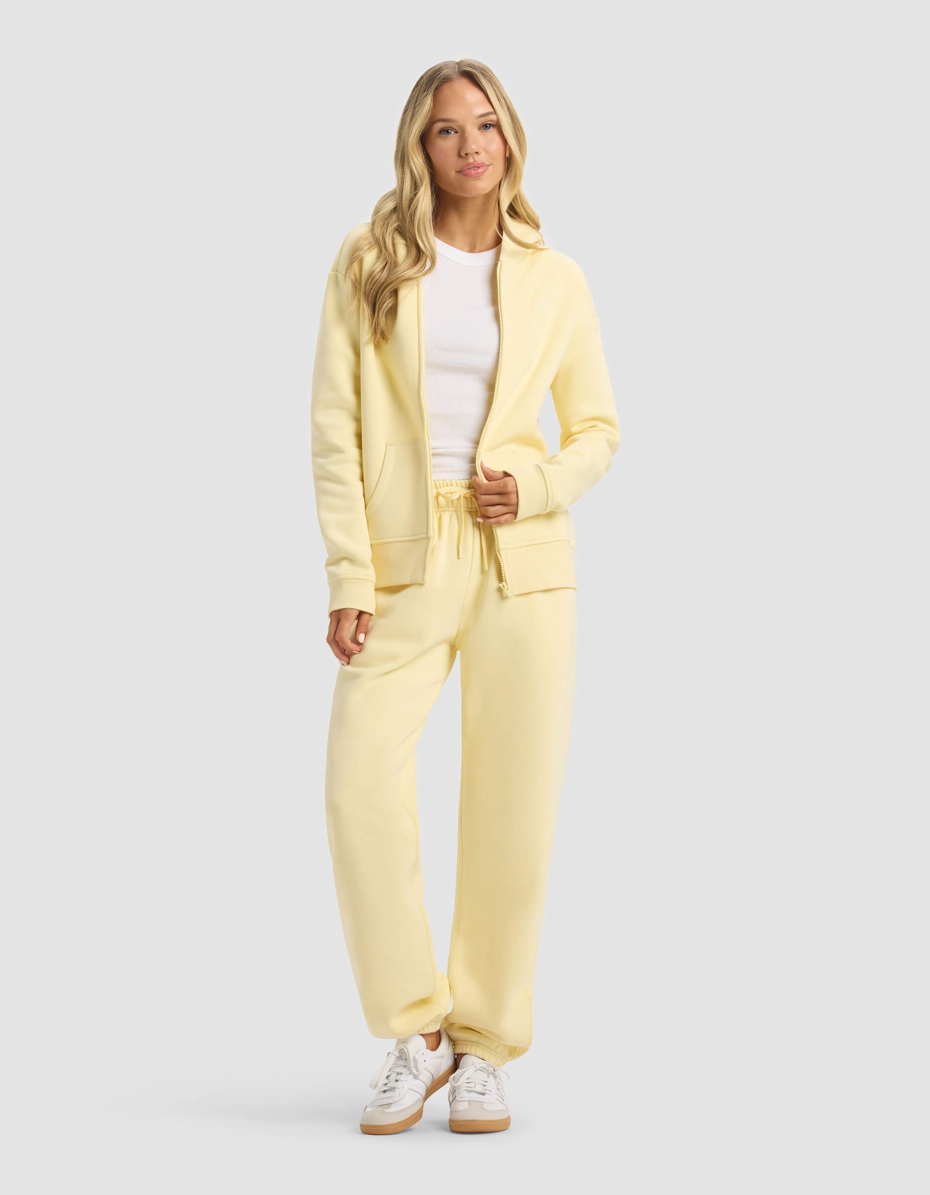 A woman with long blonde hair wears the Cozy Earth Women's CityScape 2.0 Full Zip & Sweatpant Set in light yellow, paired with a white t-shirt and sneakers. She stands facing forward against a plain light gray background.|Color:Meringue