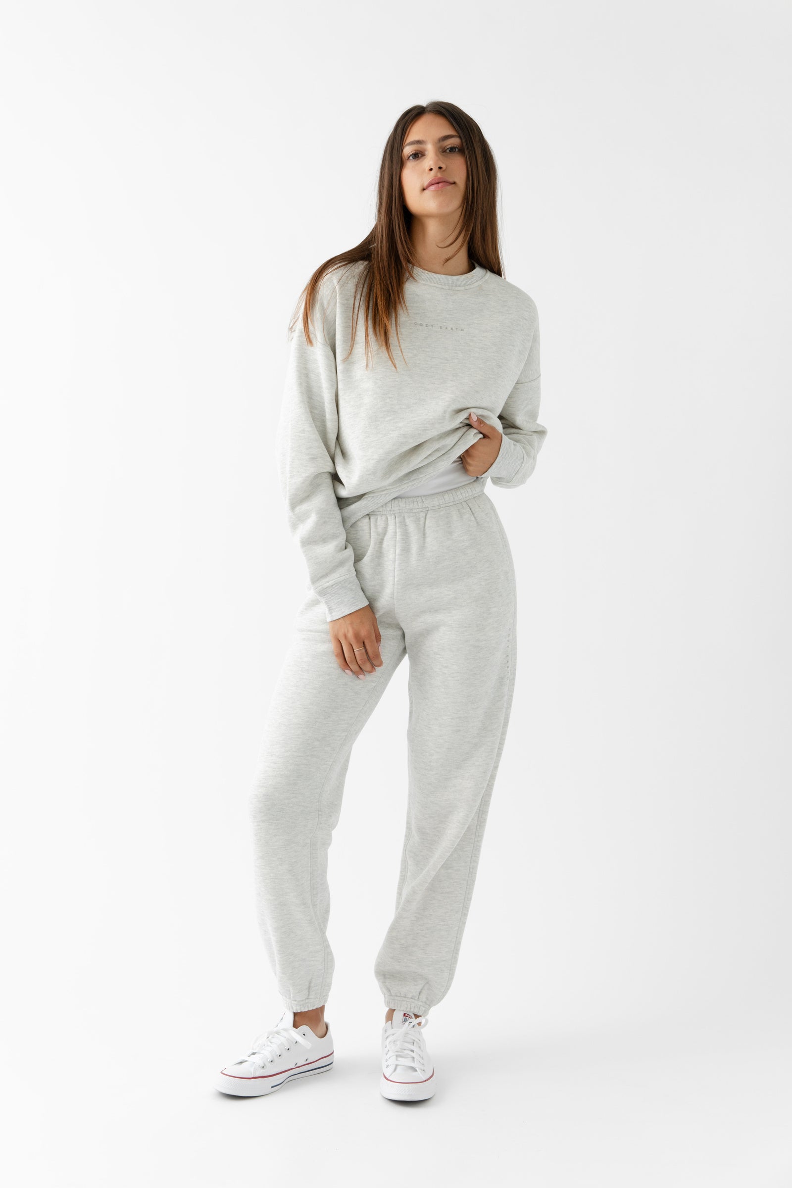 A woman with long brown hair confidently poses against a white background, wearing Cozy Earth's Women's CityScape Crewneck & Sweatpant Set in light gray with white sneakers.|Color:Heather Grey