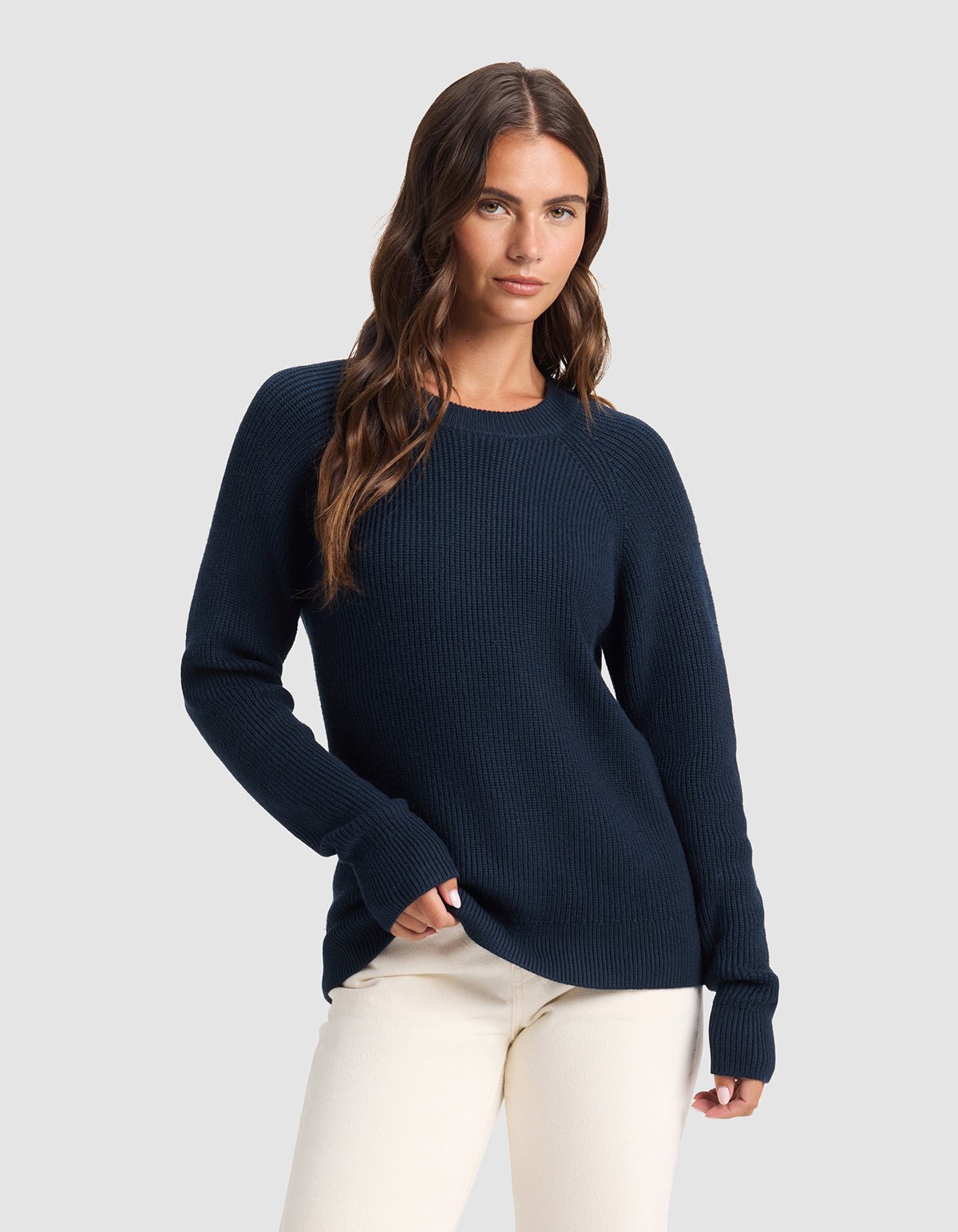 A woman with long brown hair models the Cozy Earth Women's Classic Crewneck in navy blue, paired with off-white pants. She stands against a plain light gray background, one hand in her pocket.