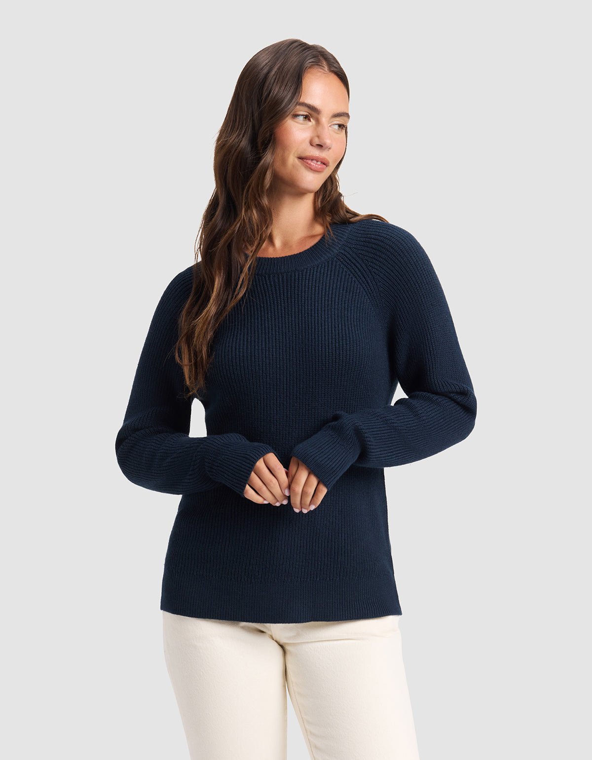 A woman with long brown hair wears the Cozy Earth Women's Classic Crewneck in navy blue and cream-colored pants, standing against a light gray background and smiling gently while looking to the side.
