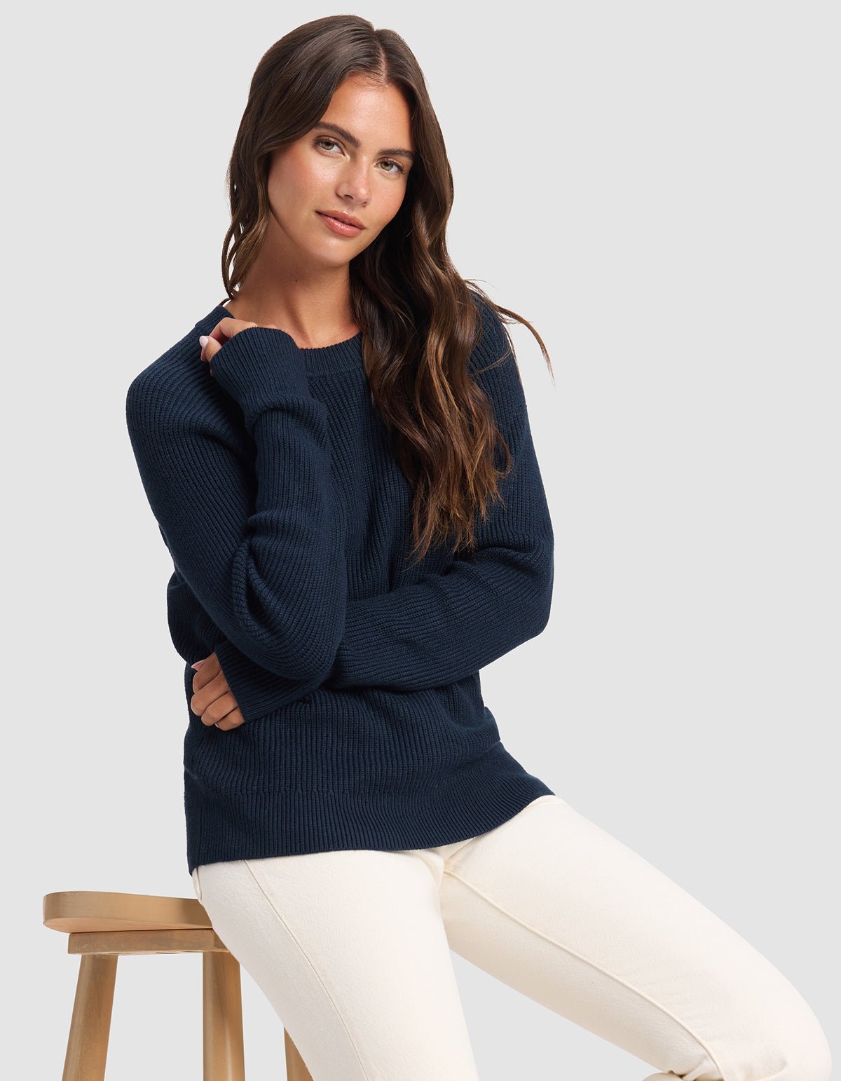 A woman with long brown hair wears the Cozy Earth Women's Classic Crewneck in navy blue and off-white pants, seated on a wooden stool against a plain light gray background.