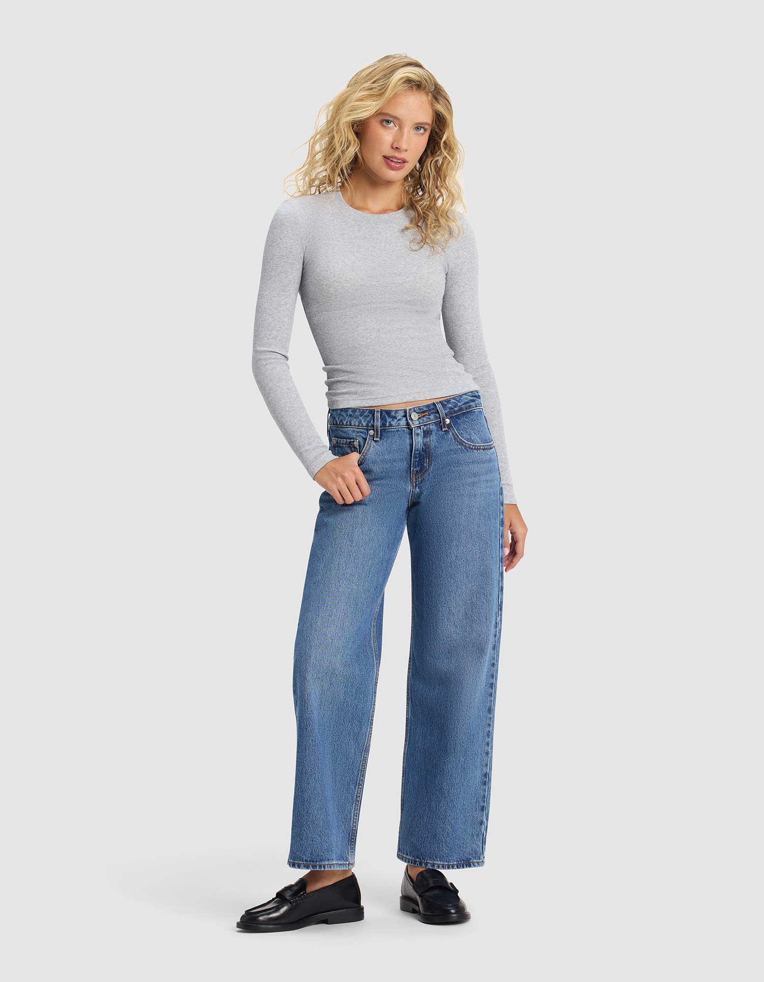 A woman with long blonde curly hair wears the Cozy Earth Women's Fine Ribbed Long Sleeve Tee, styled with blue high-waisted wide-leg jeans and black loafers, standing against a plain light background.