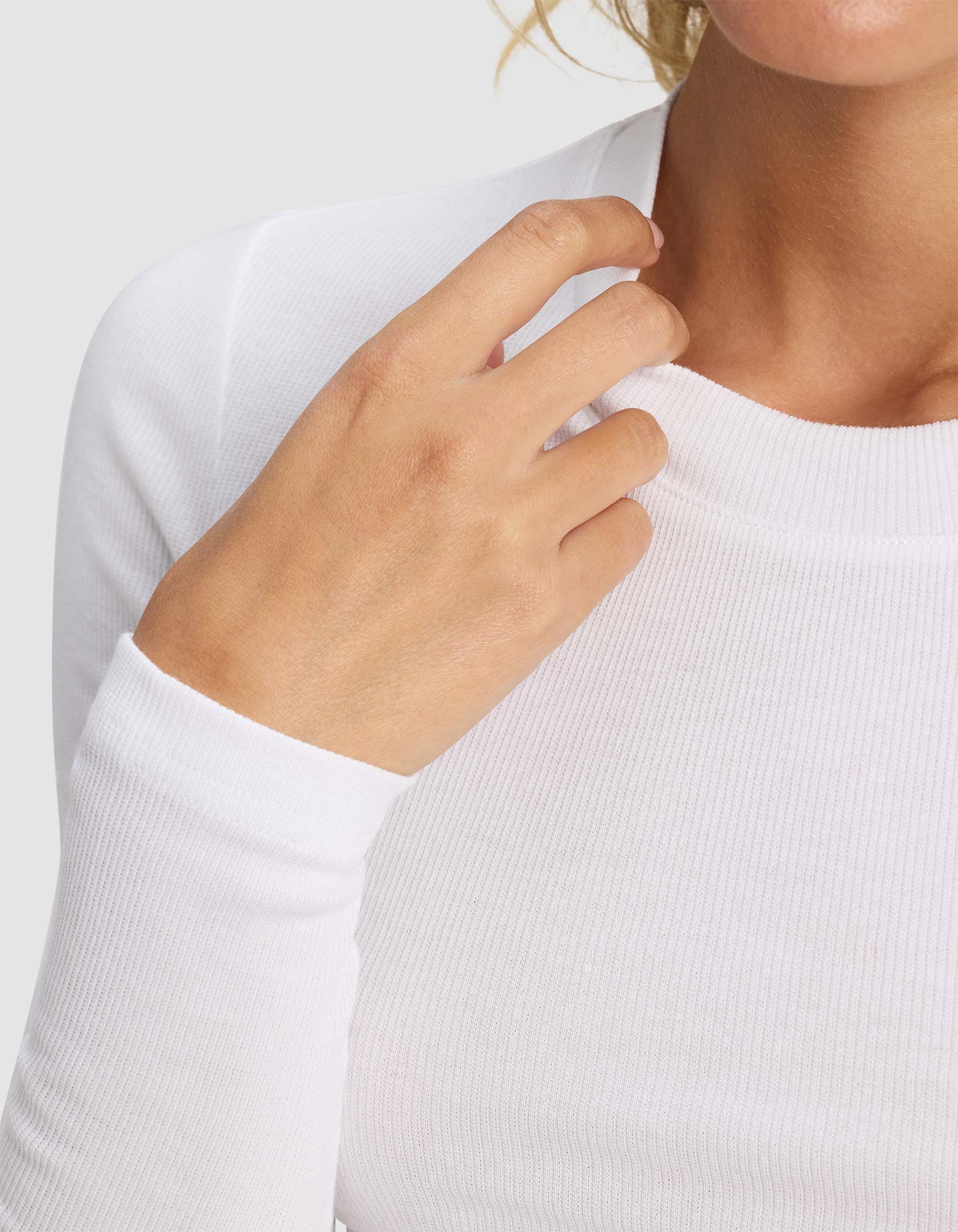 A close-up shows a person in the Cozy Earth Women's Fine Ribbed Long Sleeve Tee in white, gently touching the neckline. Their face is partly out of frame against a light gray background.