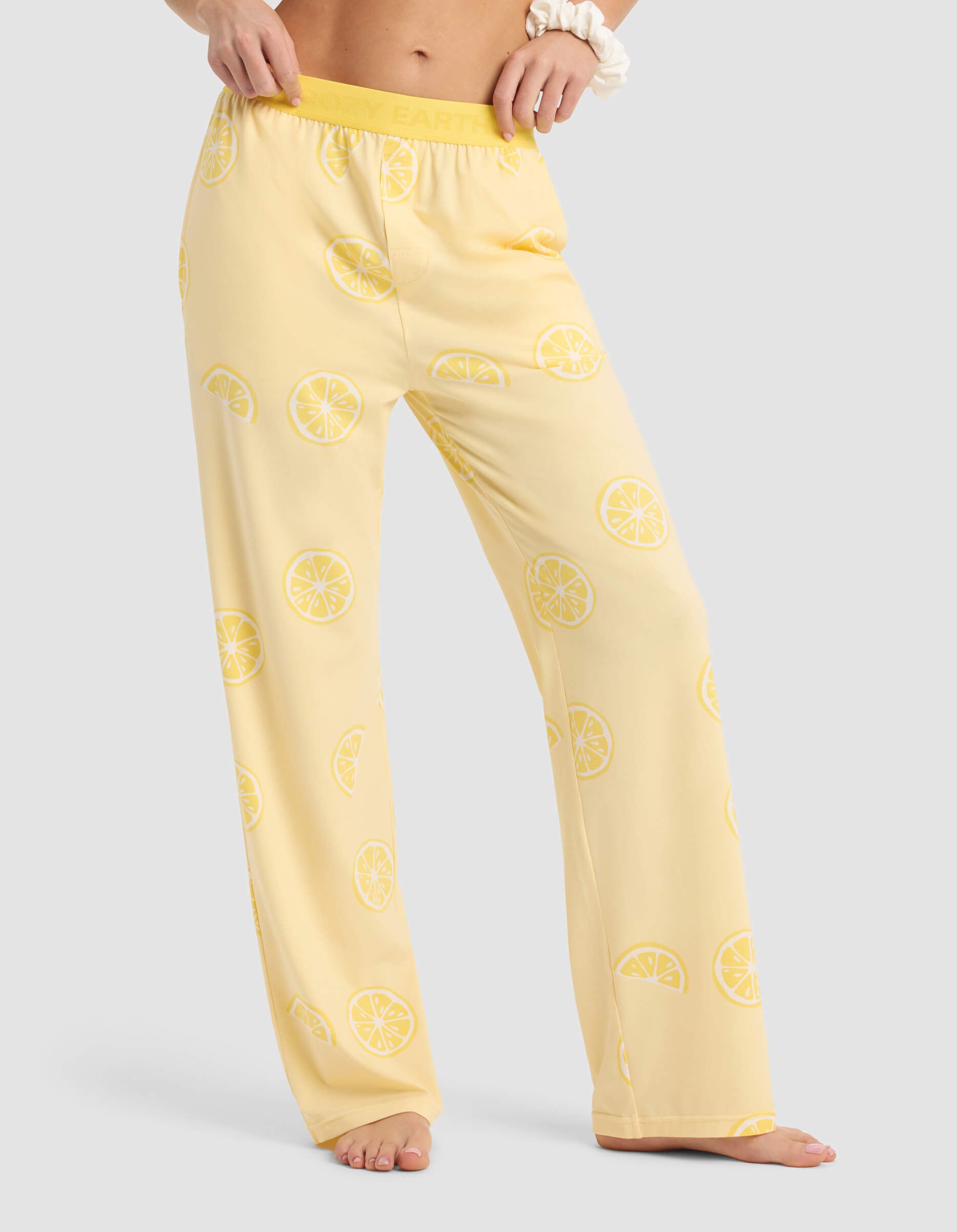 A woman wears Cozy Earth’s Women's First Layer Pajama Pant, featuring a light yellow color with lemon slice prints, a relaxed fit, and an elastic waistband, standing barefoot against a light gray background.|Color:Lemons