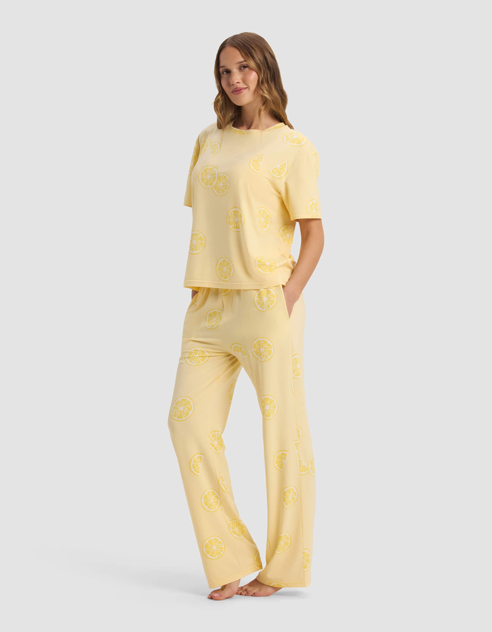 A woman stands barefoot, wearing the Cozy Earth Women's First Layer Pajama Short Sleeve Tee & Pant Set in light yellow, featuring a loose fit and an all-over yellow lemon slice print.|Color:Lemons