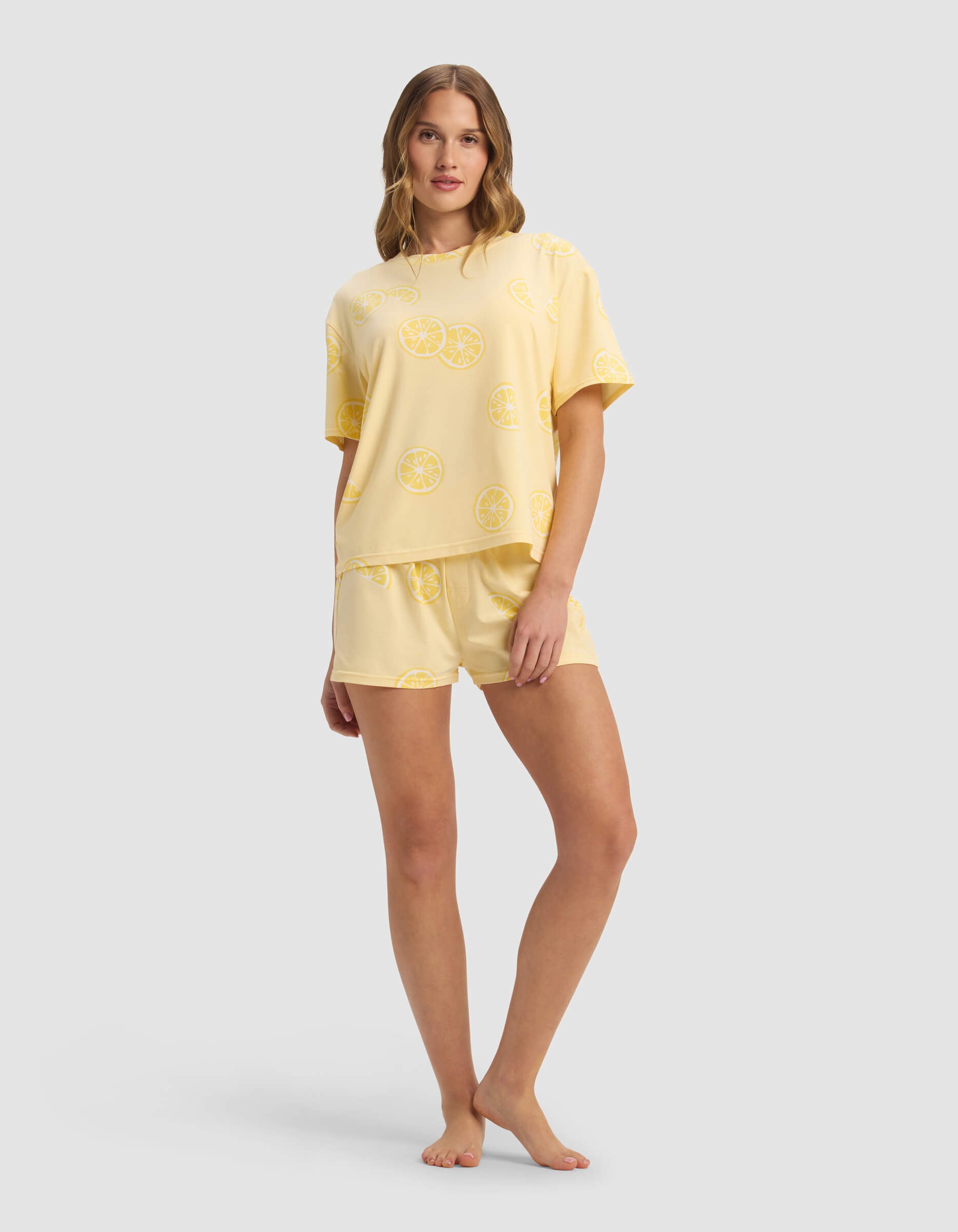 A woman stands barefoot against a plain light gray background, wearing the Cozy Earth Women's First Layer Pajama Short Sleeve Tee & Boxer Short Set in a matching yellow citrus slice pattern.|Color:Lemons