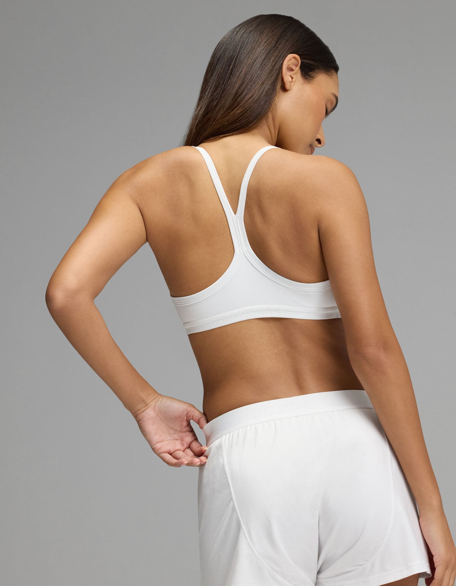 A woman stands with her back to the camera, wearing the Cozy Earth Women’s First Layer Soft Bra in white, paired with matching white shorts, against a plain gray background.|Color:White