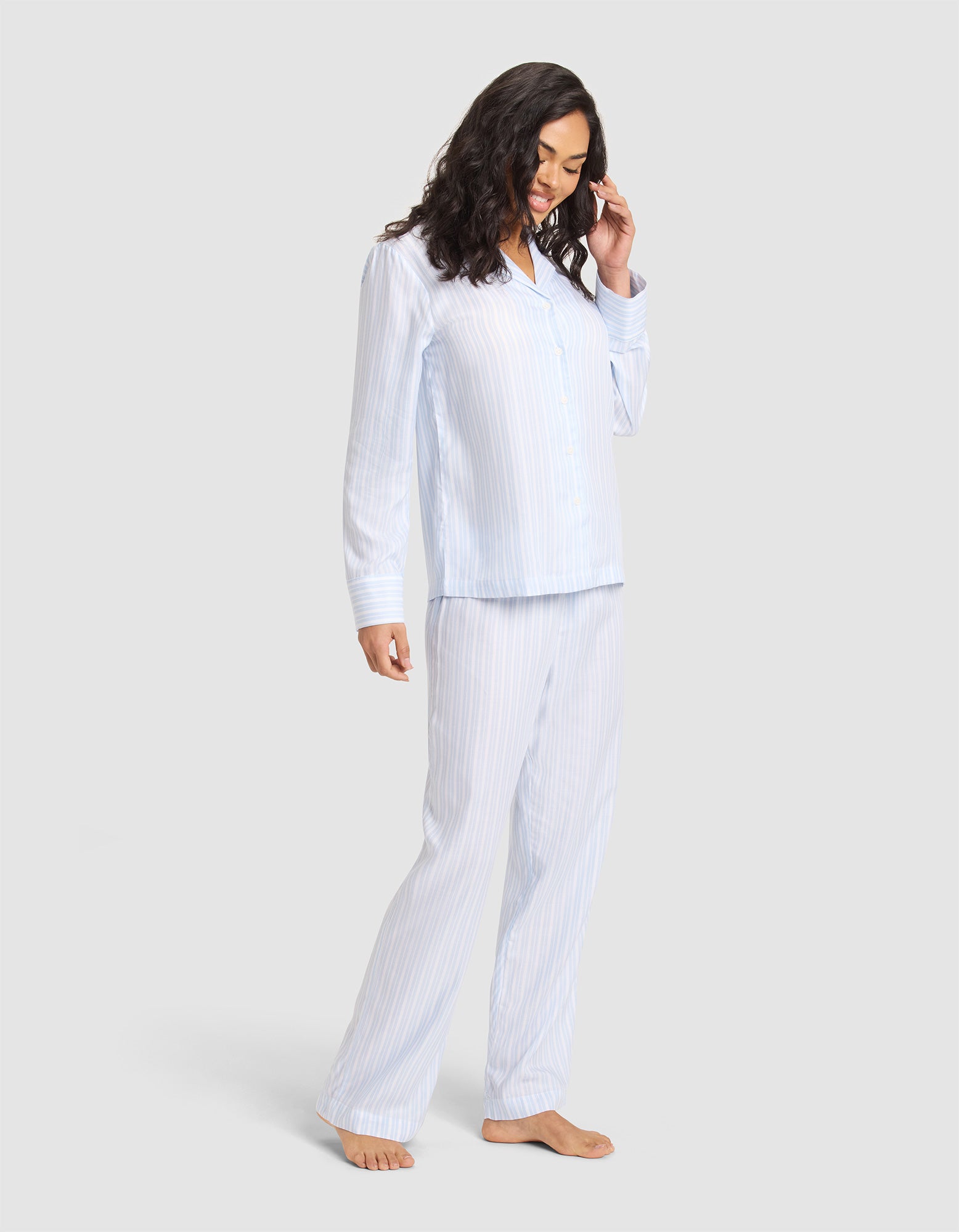 A woman with long dark hair, barefoot and smiling, models the Cozy Earth Women's Soft Woven Long Sleeve Pajama Set—light blue and white striped—against a plain light gray background.