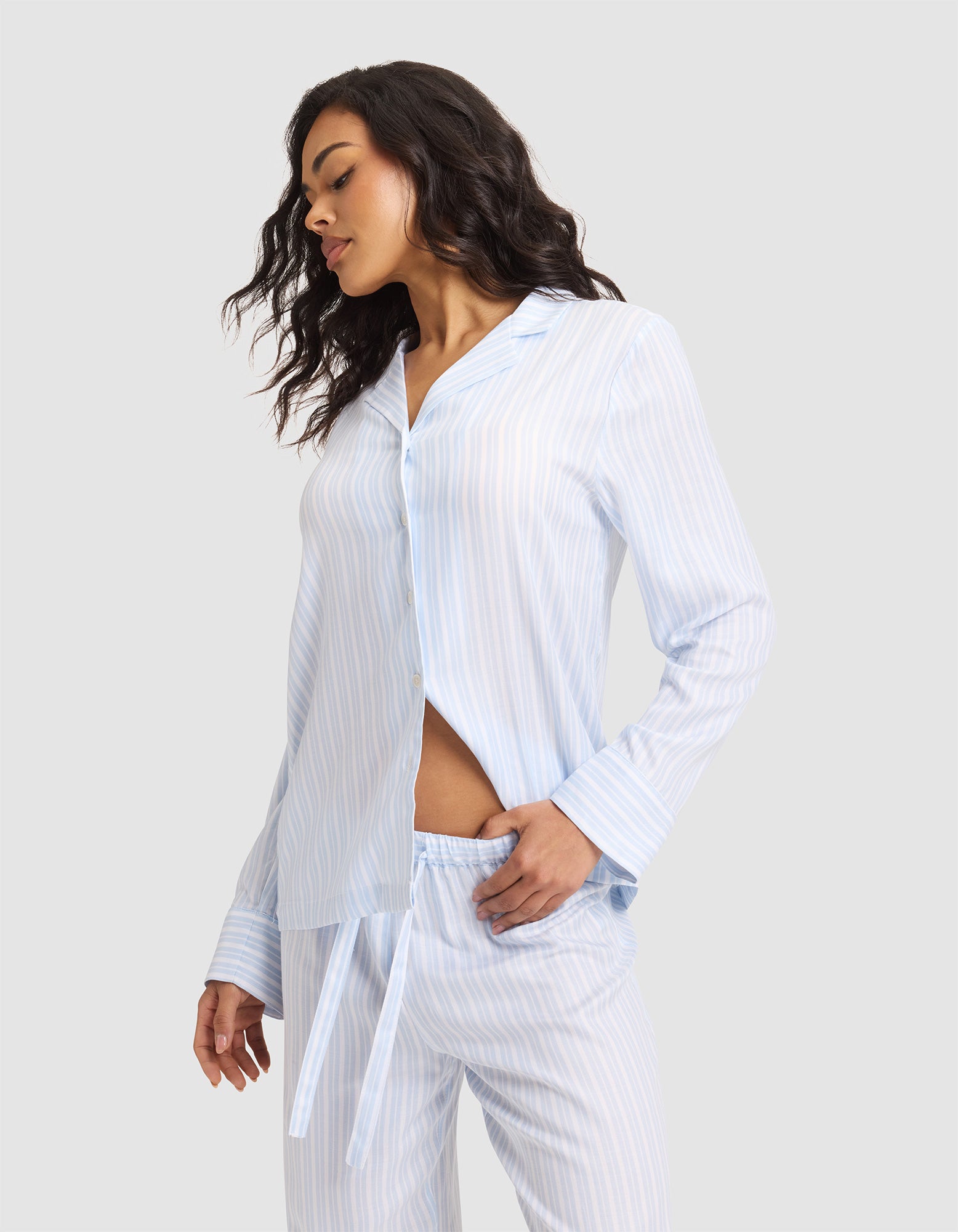 A woman stands sideways in a Cozy Earth Women's Soft Woven Long Sleeve Pajama Top, light blue and white striped, with buttons and matching pants, one hand in her pocket against a plain light background. |Color:Spring Blue Stripe