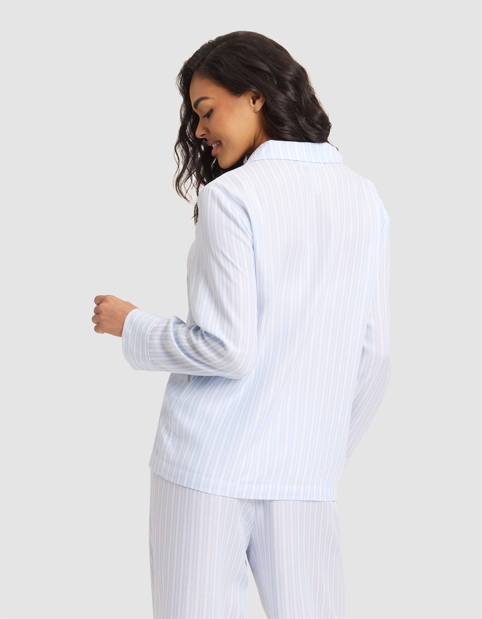A woman with wavy hair smiles and looks down, shown from the back and slightly turned to the side, wearing the Cozy Earth Women's Soft Woven Long Sleeve Pajama Top in light blue and white stripes against a plain light gray background.