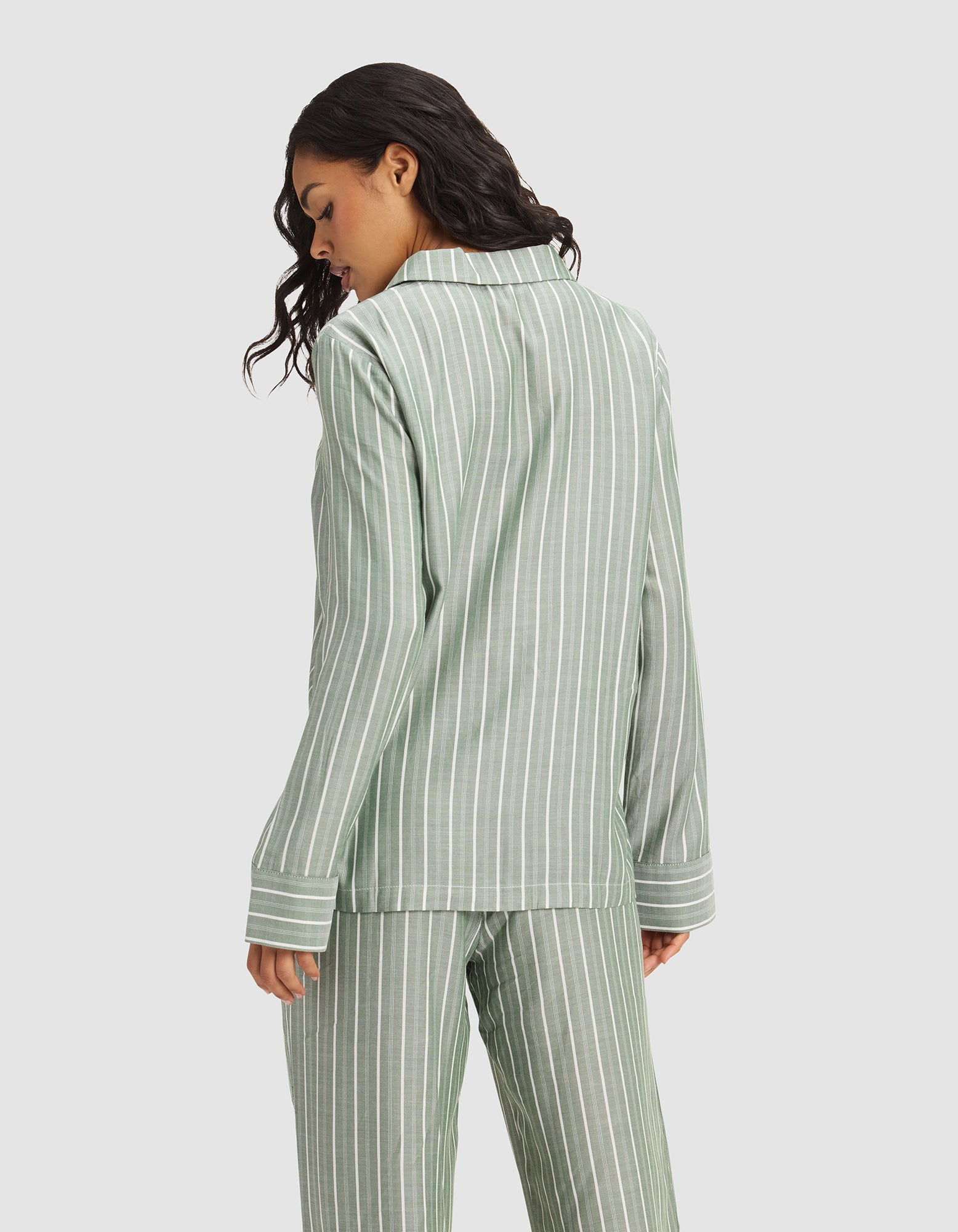 A woman with wavy dark hair models the Cozy Earth Women's Soft Woven Long Sleeve Pajama Top in sage green with white stripes, shown from the back against a plain light gray background.