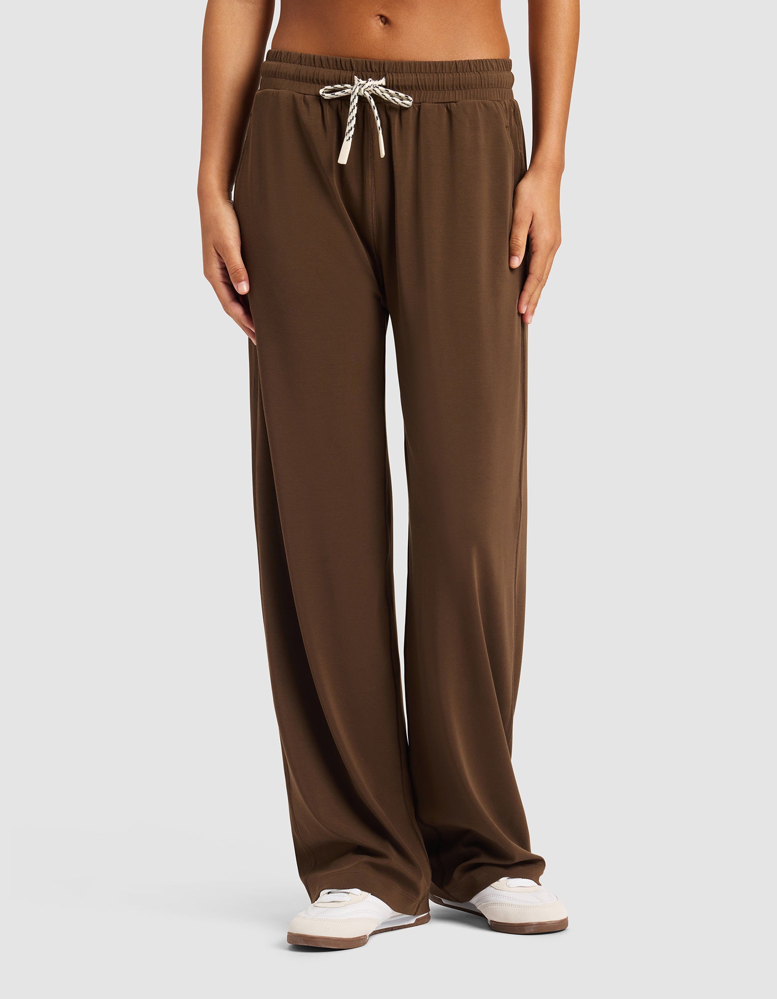 A person is standing against a light gray background wearing Cozy Earth's Women's Studio Wide Leg Pant in brown with white sneakers, hands relaxed by their sides. The pants feature a loose fit and drawstring waist.|Color:Espresso