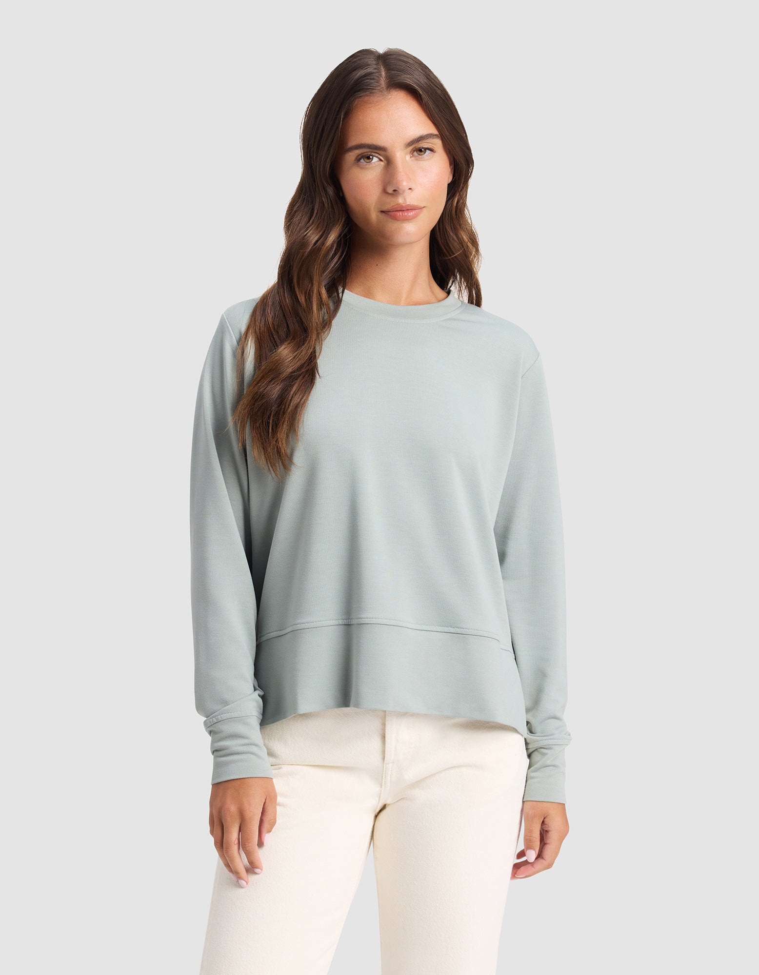 A woman with long brown hair wears the Cozy Earth Women's StudioLite Crewneck in light green and cream-colored pants, standing against a plain light gray background.|Color:Haze