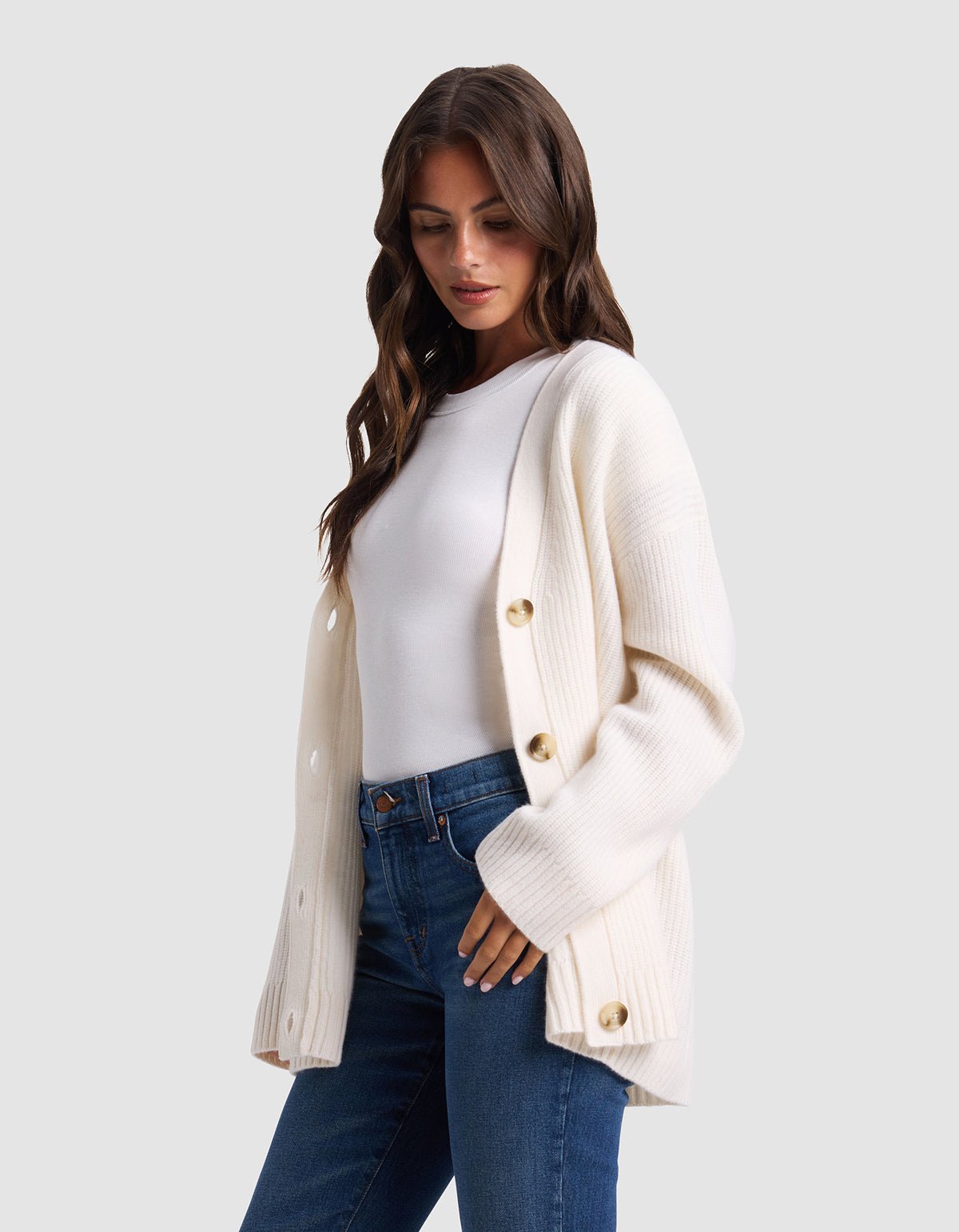 A woman with long brown hair wears the Cozy Earth Oversized Sunday Cashmere Cardigan in white over a white top and blue jeans, standing against a plain light gray background.