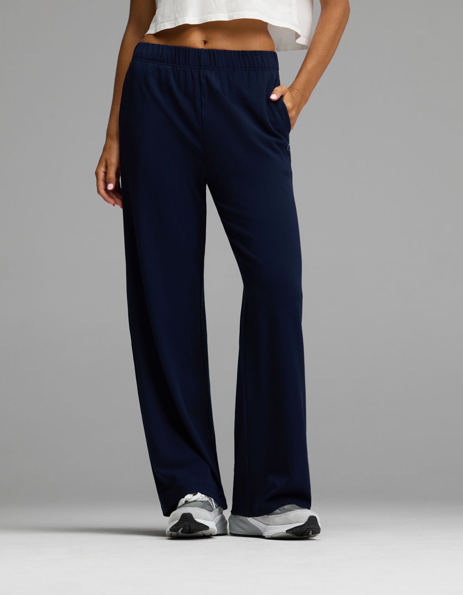 A person wearing the Cozy Earth Women’s Brushed Bamboo Wide Leg Pull-On Pant in Navy, paired with a white cropped top and white sneakers, stands with one hand in their pocket against a plain gray background.|Color:Navy