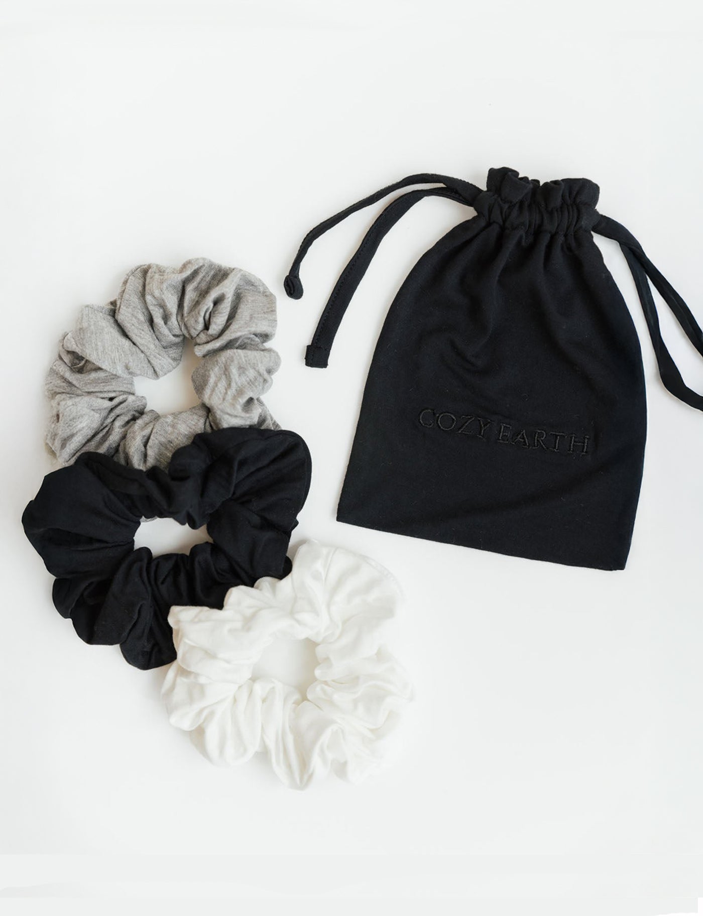 Three Bamboo Stretch-Knit Scrunchies by Cozy Earth in gray, black, and white are displayed beside a black Cozy Earth drawstring pouch against a white background.|Color:Black/Ivory/Grey