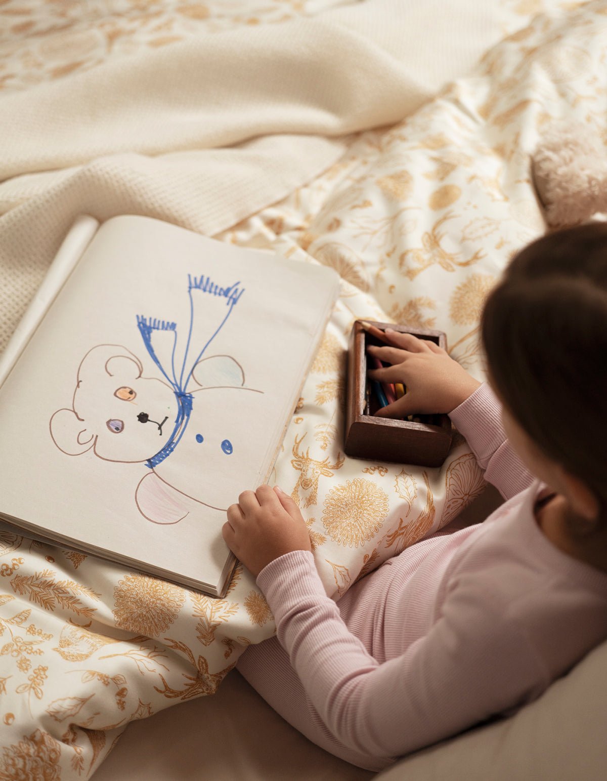 A child in pink pajamas sits on a bed made with Cozy Earth’s Bamboo Sheet Set, drawing a bear in a scarf with crayons in a sketchbook, while their hand rests on a box of crayons.|Color:Winter Toile Dijon