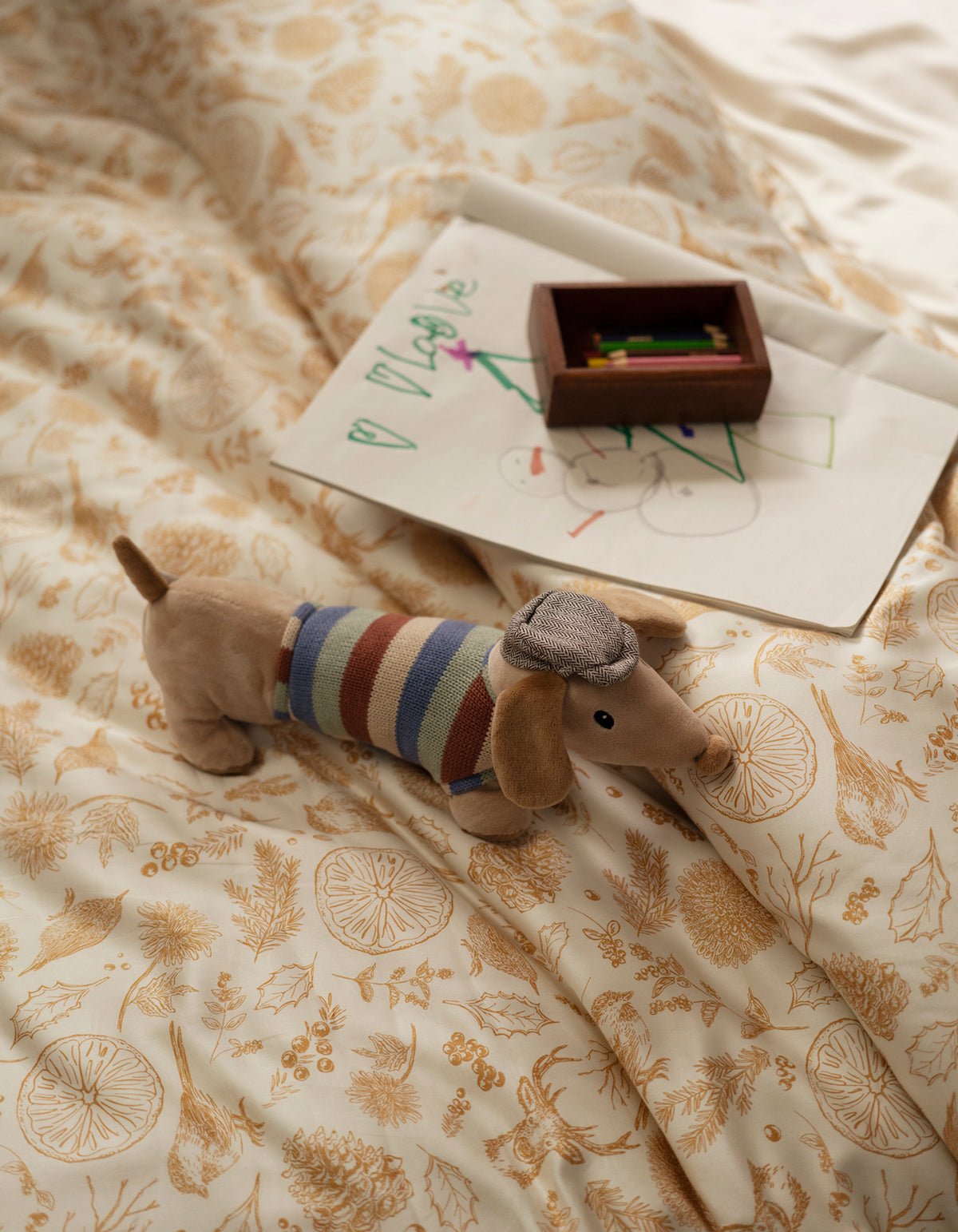 A plush dachshund toy in a striped sweater and hat rests on a bed made with the Cozy Earth Bamboo Duvet Cover; colored pencils and a child’s drawing on white paper are nearby.