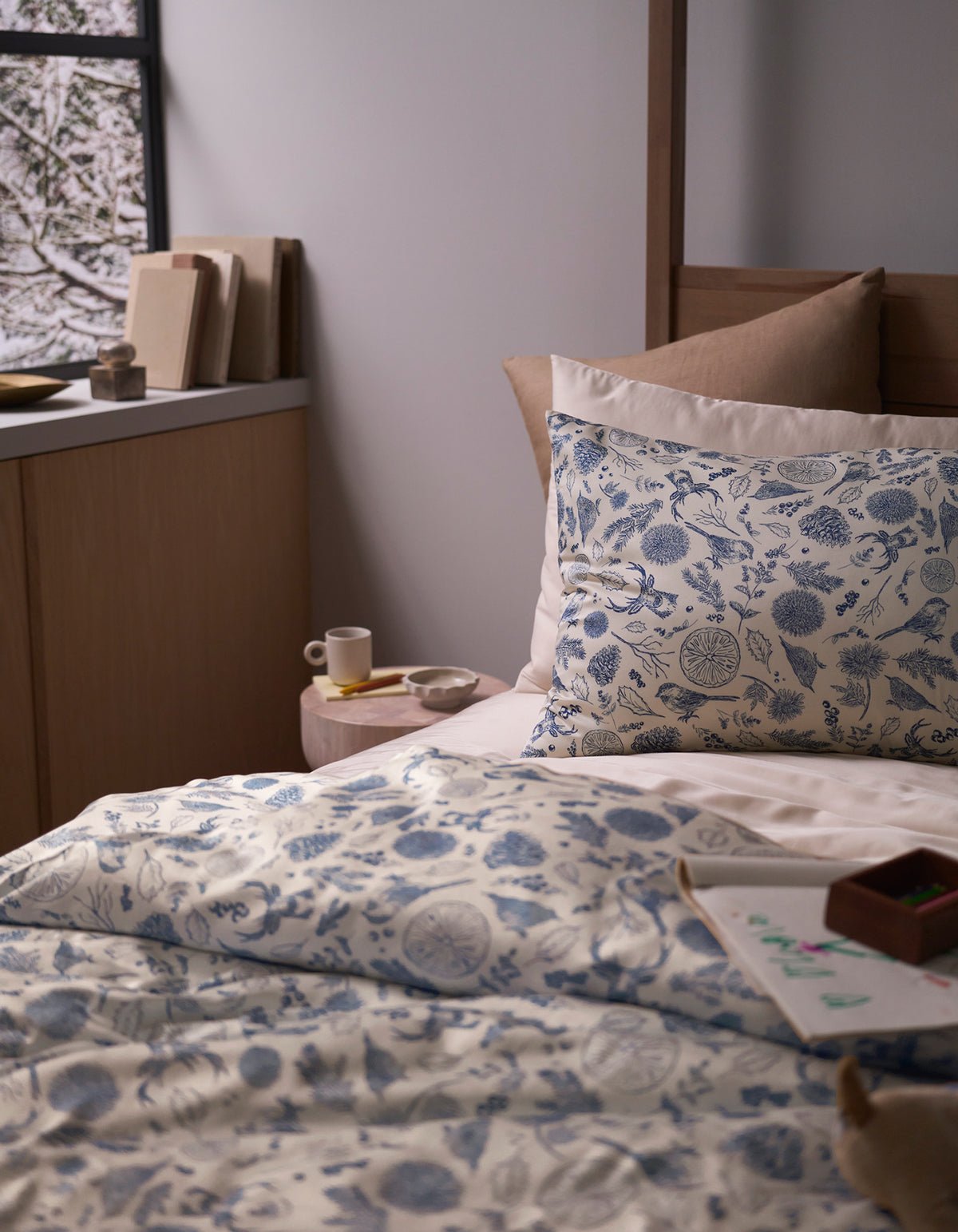 A cozy bedroom features a bed topped with the Cozy Earth Bamboo Sheet Set, beige pillows, a small bedside table holding a cup and saucer, books, and a window displaying a snowy scene outside.|Color:Winter Toile Pacific