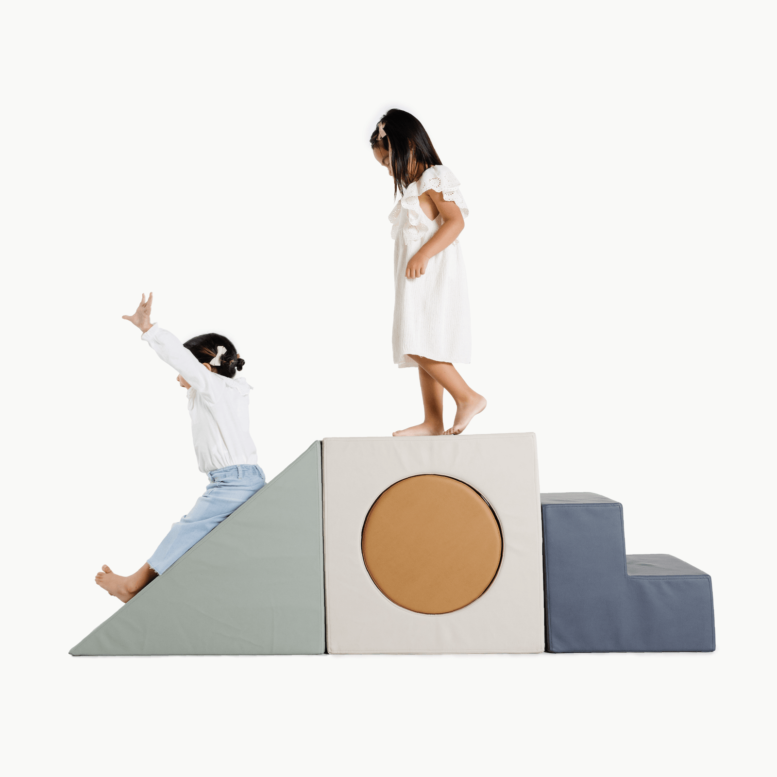 Two young girls enjoy the Gathre Wholesale Block Playset against a white background—one slides down the ramp, while the other stands atop the soft foam structure with a ramp, cube with a circle cutout, and steps.